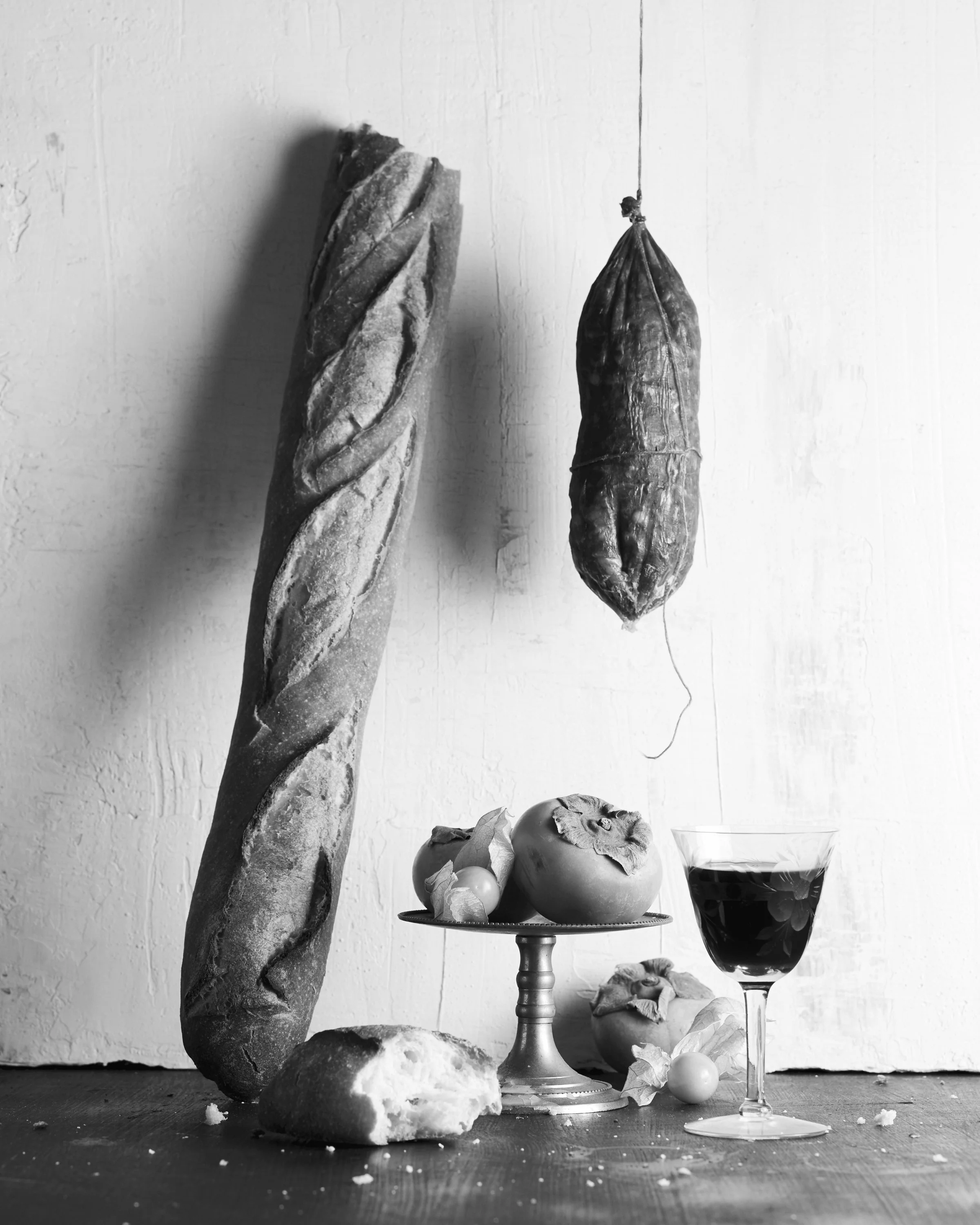 Black and white image featuring a baguette, hanging sausage, glass of red wine, and fruits on a pedestal.