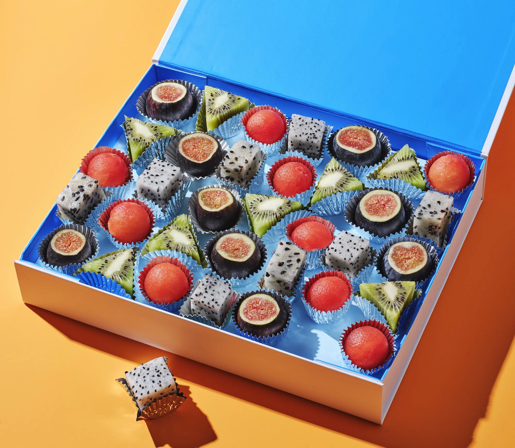 Nature’s Candy is a collaborative series by photographer Sarah Anne Ward & food stylist Charlotte Omnes A candy box of assorted exotic fruit pieces, including kiwi, figs, and dragon fruit, arranged in colorful paper cups on a blue background.