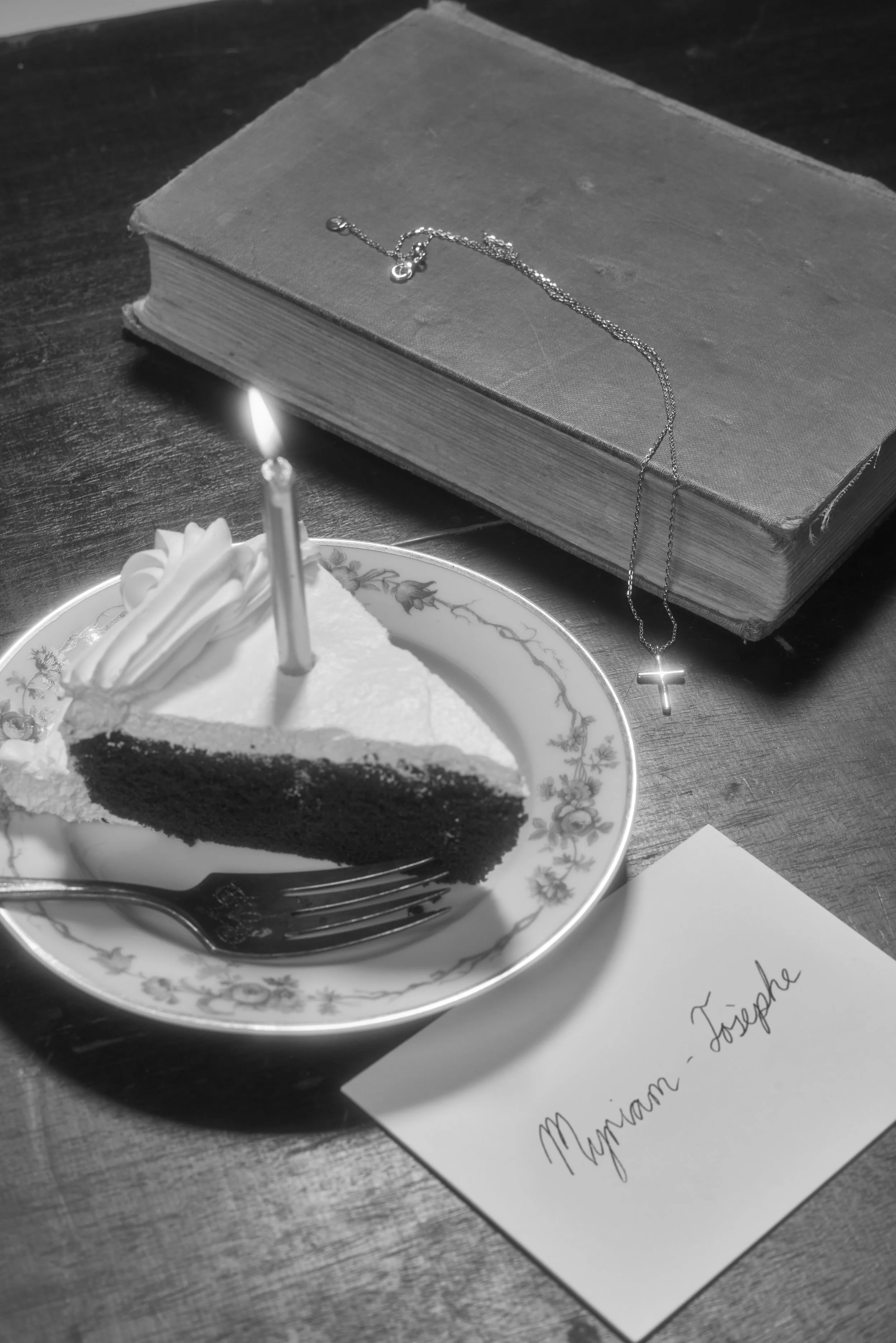 Slice of cake with candle on floral plate, old book with necklace, and envelope on wooden table.