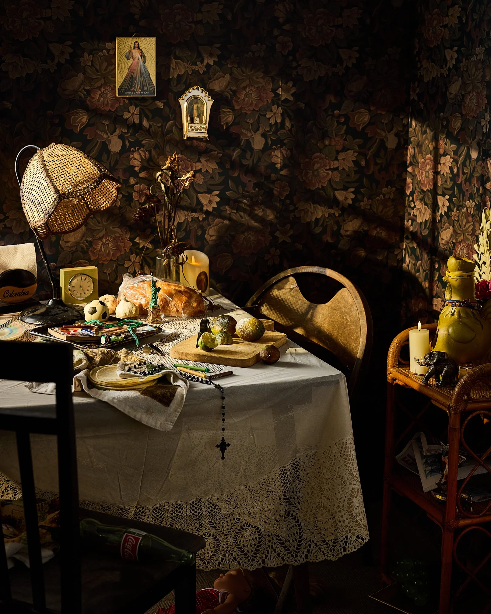 A cluttered dining table with a lace tablecloth, topped with various objects including a black and white soccer ball, a dinner roll, a small replica of the Statue of Liberty, and some fruits. The background features a dark floral wallpaper, with wall decorations including religious images, a gold lamp, and a small clock. A person wearing a red dress is seen partially under the table, with only their head and legs visible, and there is a bottle of Coca-Cola on a piece of furniture nearby.