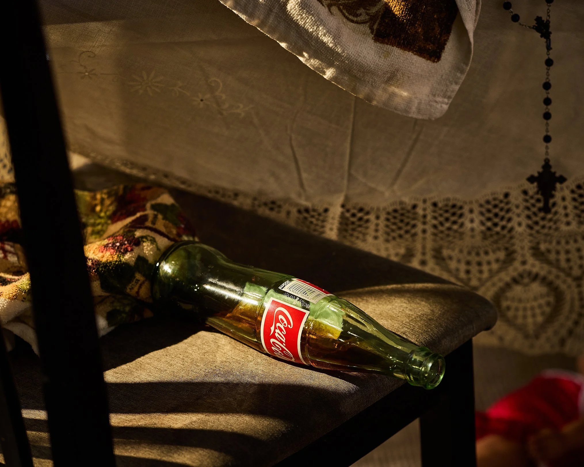 A clear glass Budweiser beer bottle lying on its side on a fabric surface, with a partially visible colorful patterned cloth nearby and a dark background.