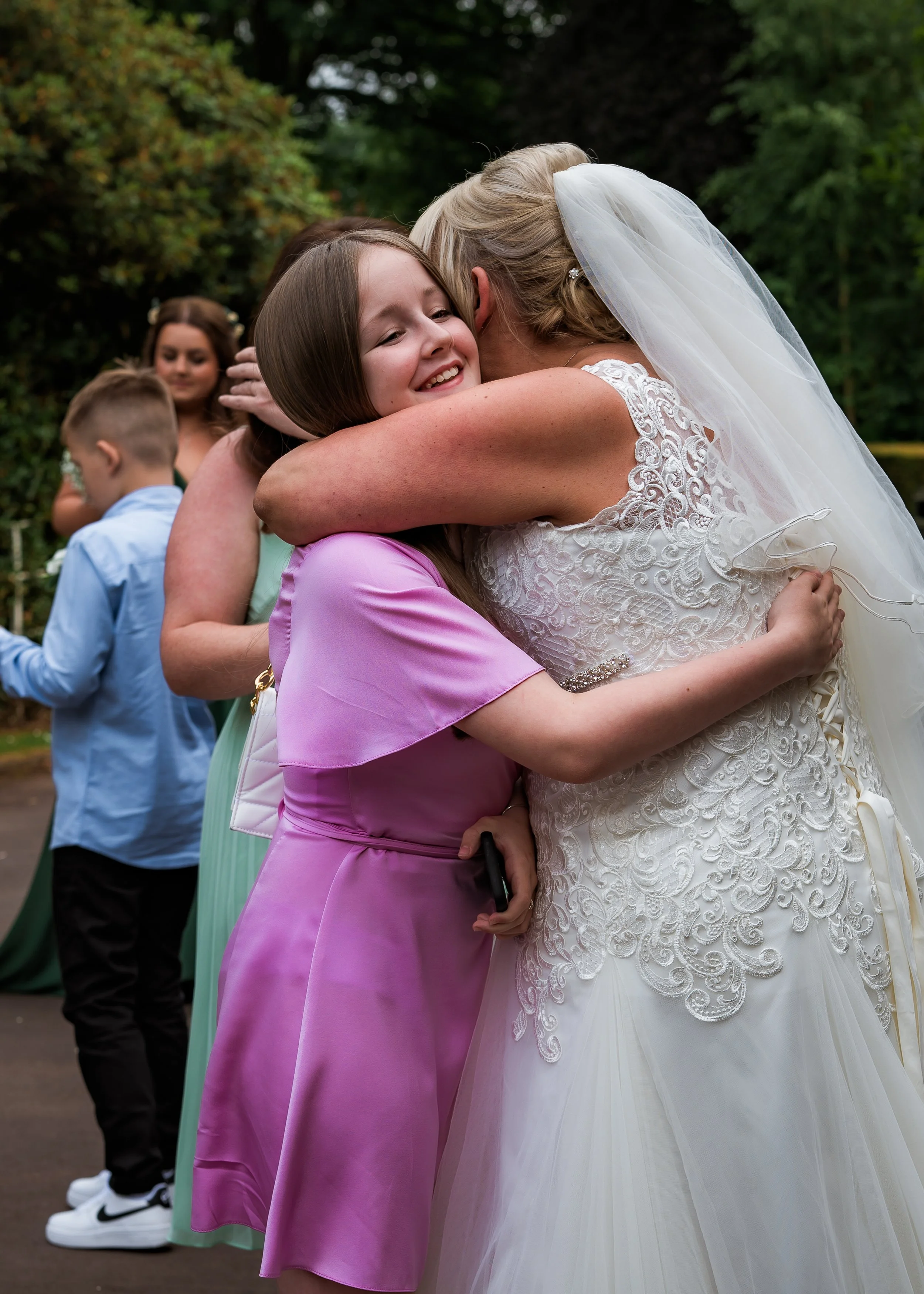 A bride in a white wedding dress and veil hugging a young girl in a pink dress at an outdoor wedding reception, with other guests in the background.