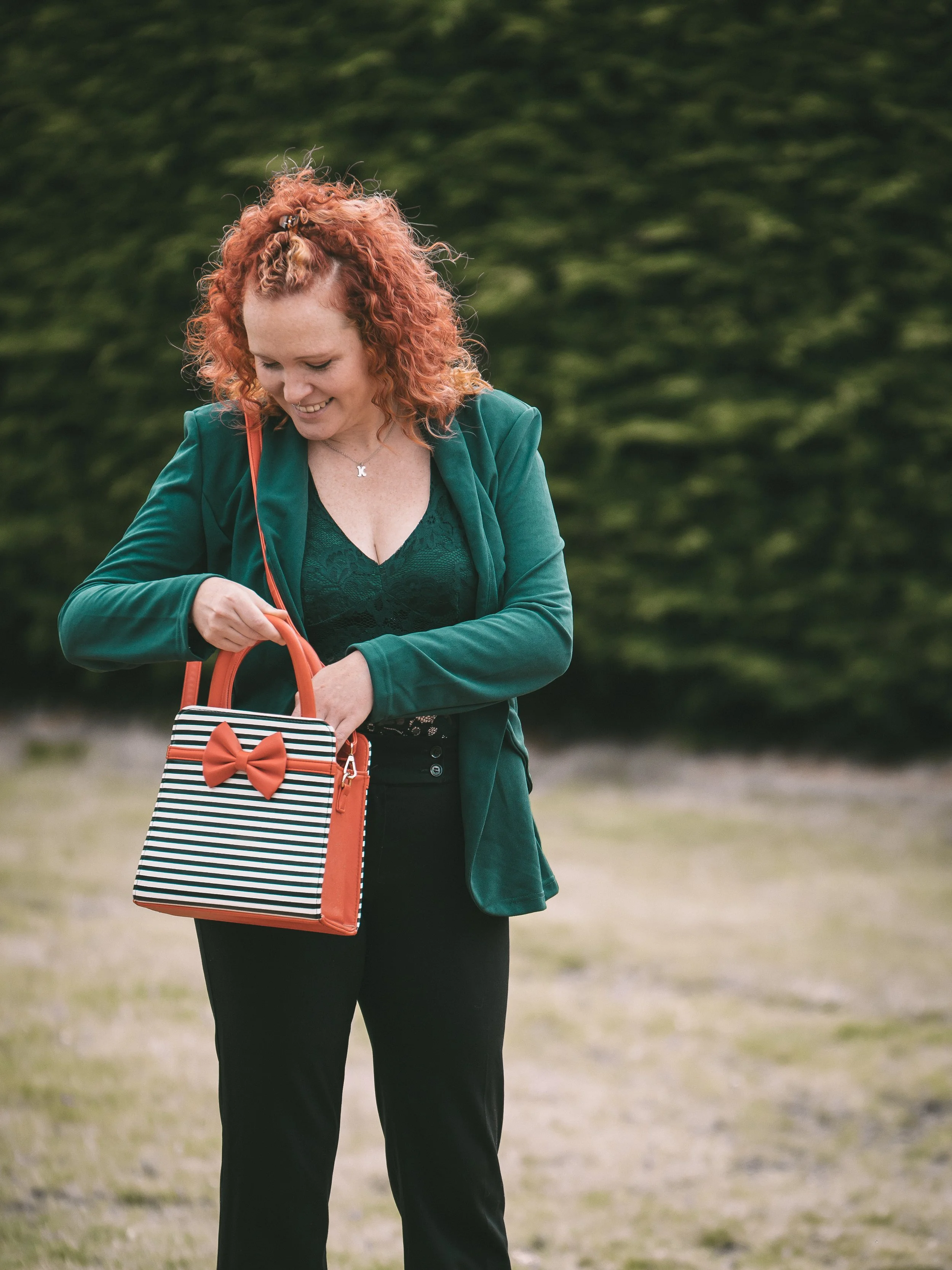 A woman with curly red hair, wearing a teal blazer and black pants, smiling while looking into her orange and black striped handbag with a bow on it, outdoors with a background of blurred green trees.