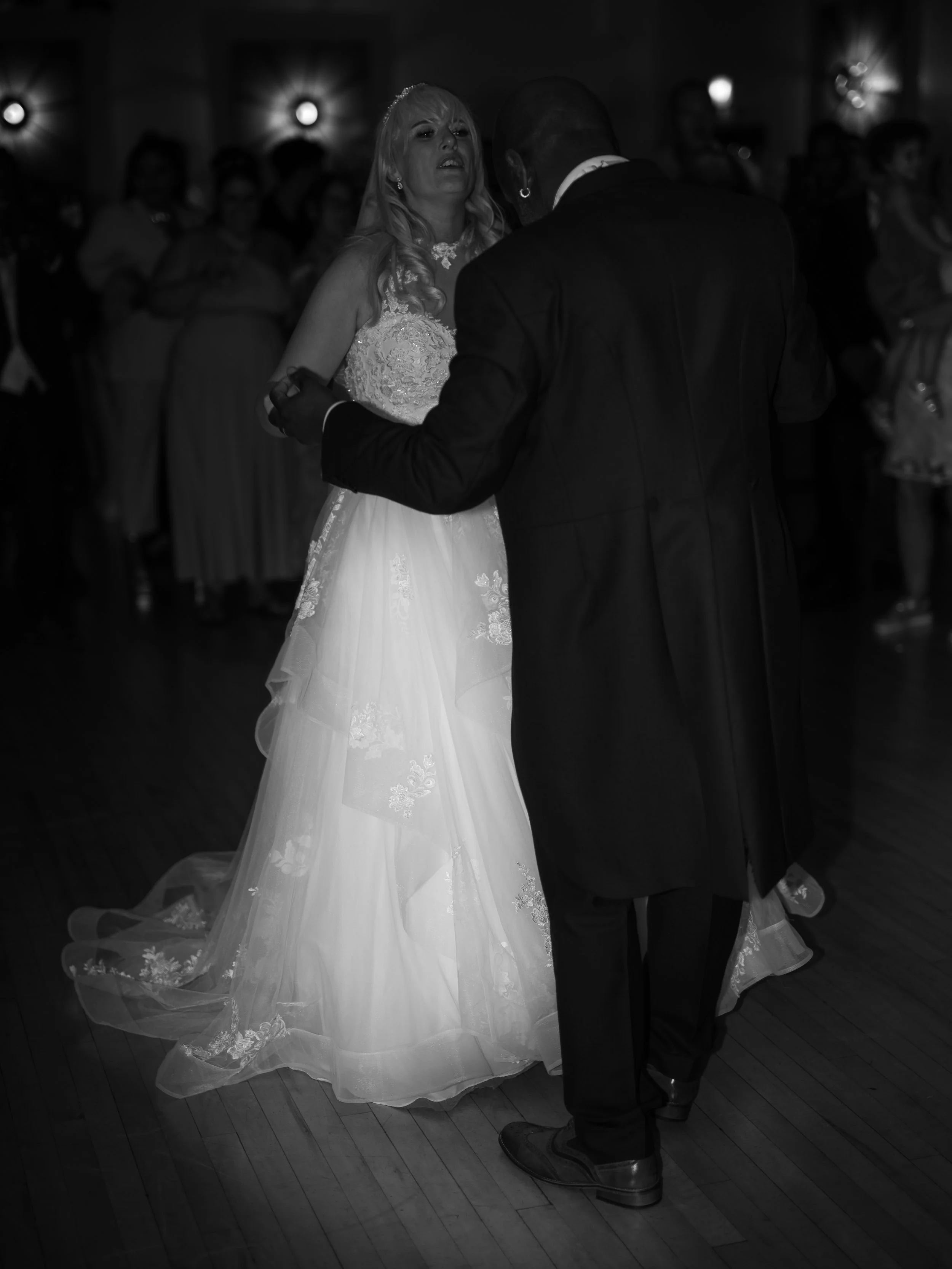 A bride and groom dancing at their wedding reception, with wedding guests watching in the background.