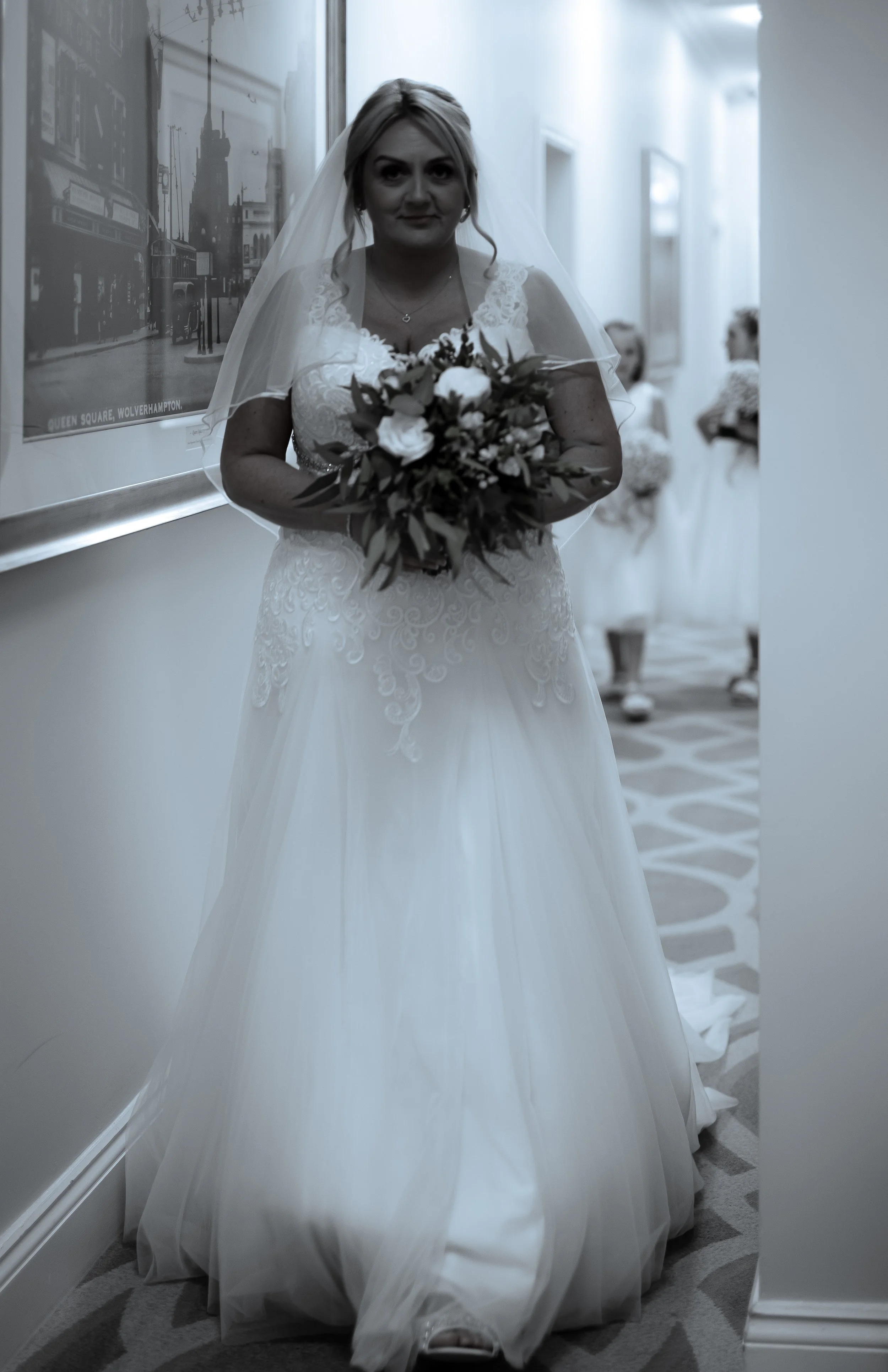 A bride in a white wedding dress and veil holding a bouquet of flowers standing in a hallway.