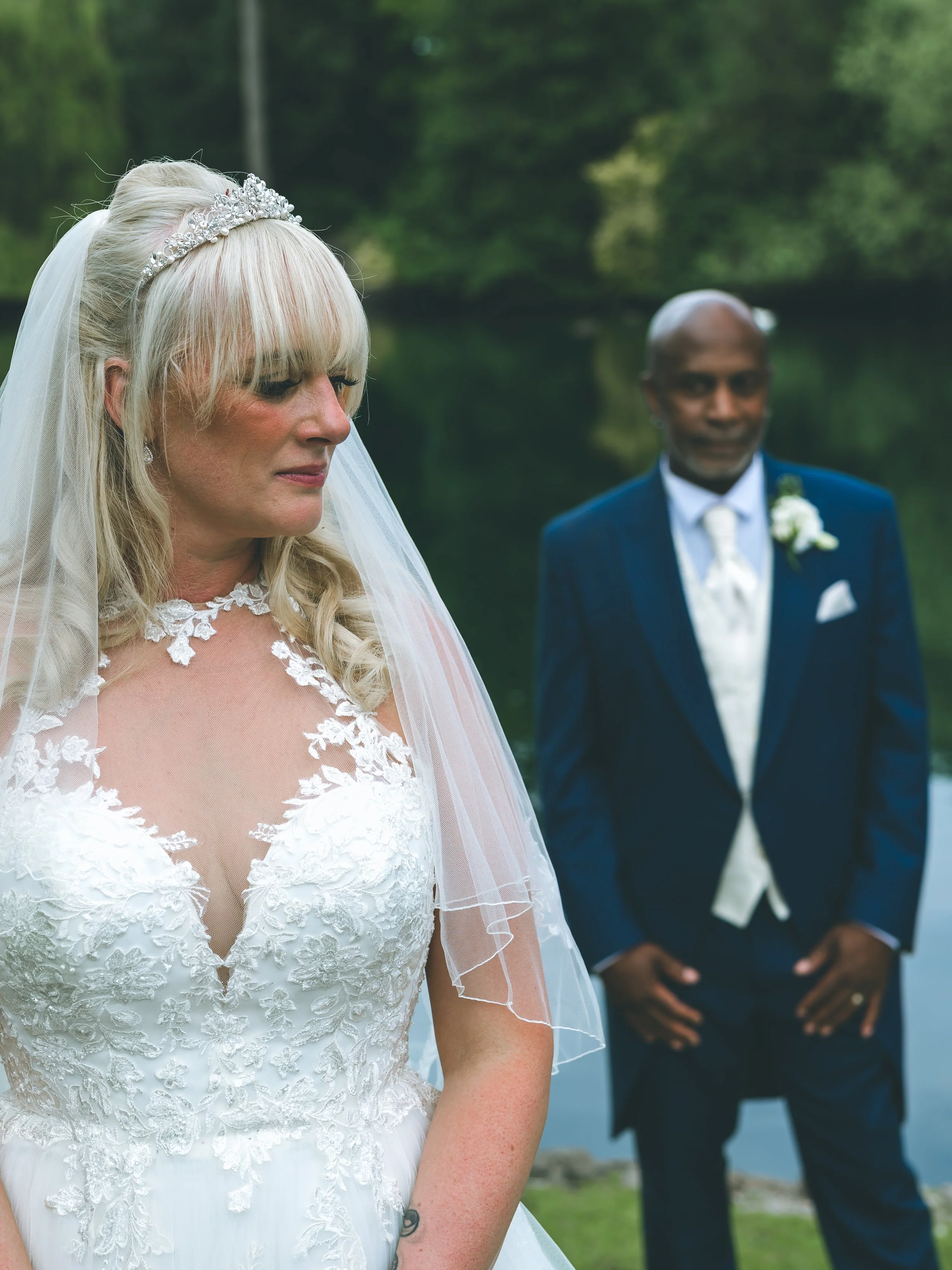 A bride in a white wedding dress with lace detailing and a veil, standing outdoors with a groom in a navy suit and white shirt blurred in the background. The bride has platinum blonde hair and a tiara, with a natural green and lake scenery behind the