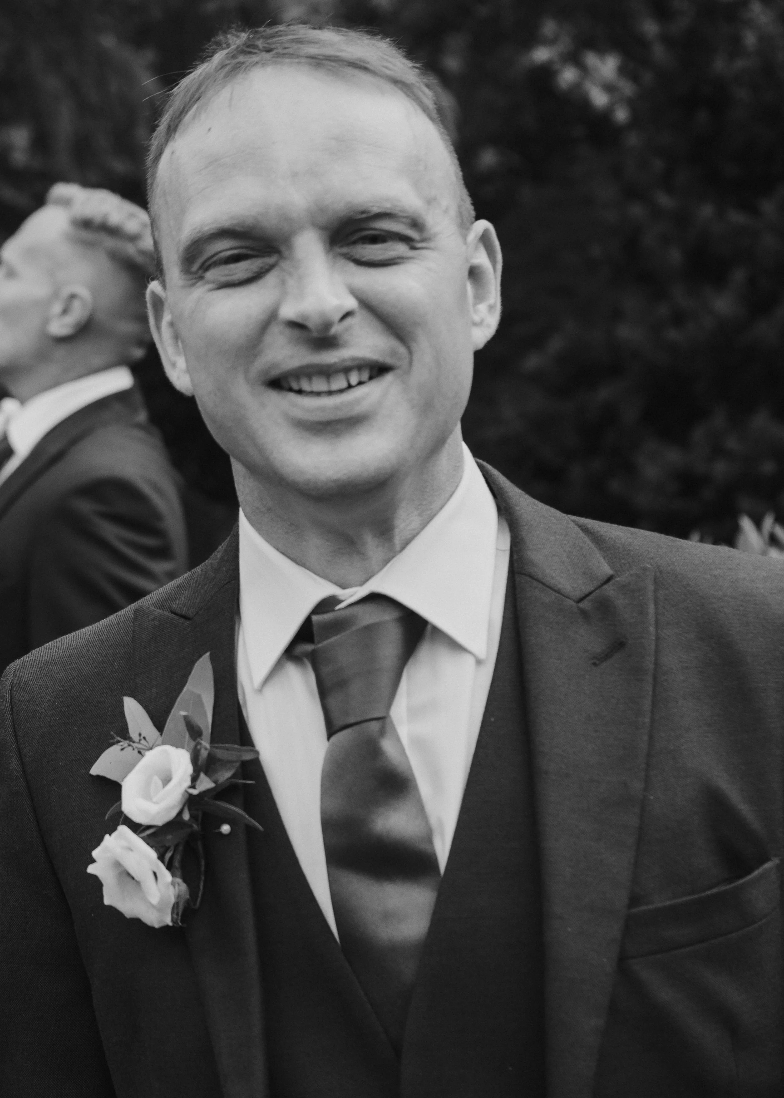 Black and white photograph of a smiling man in a suit, tie, and boutonniere, at a formal event or wedding, with his head slightly tilted and another person in the background.