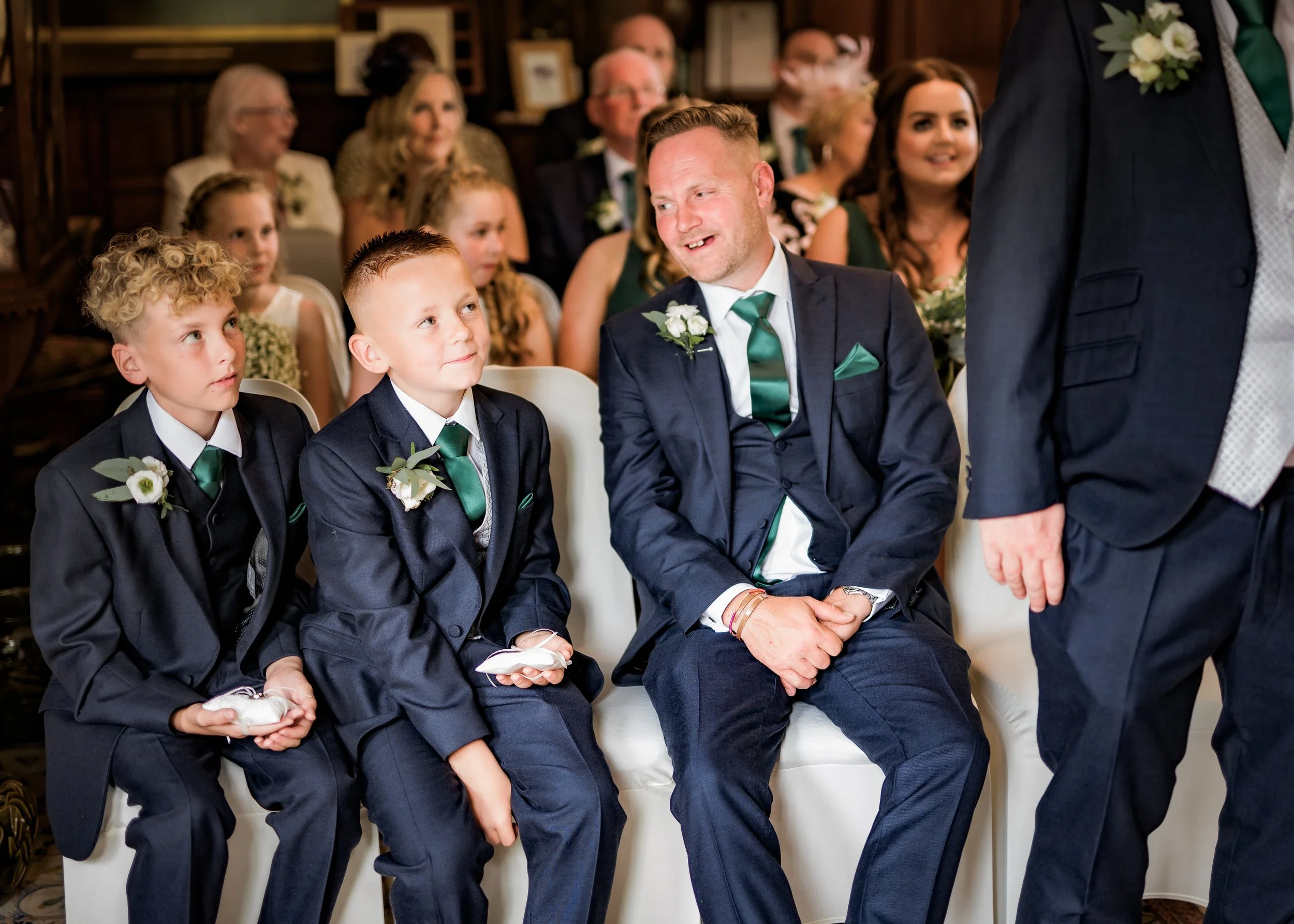 A wedding ceremony with two young boys in formal suits and three men in suits, seated indoors with a group of adults in the background.