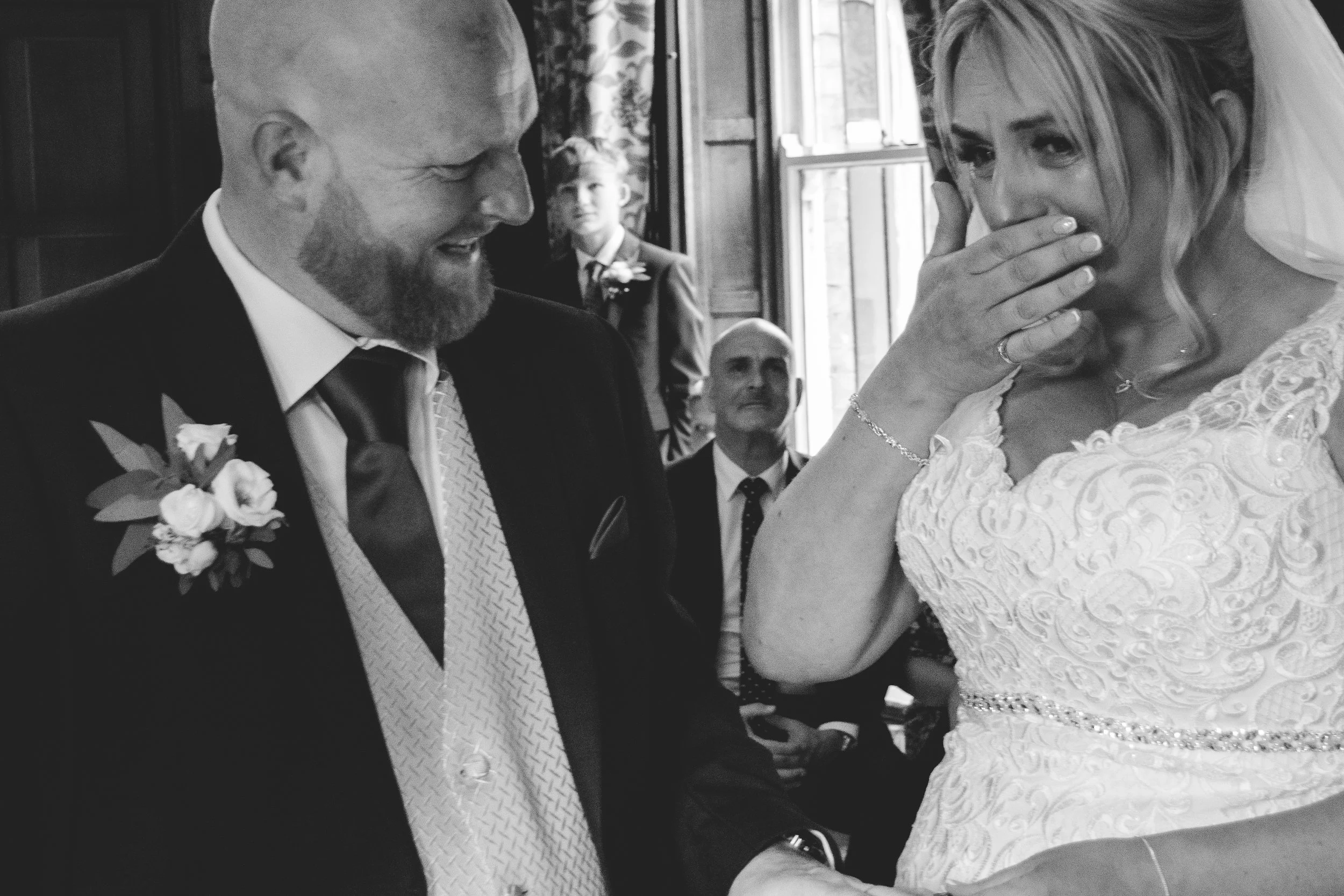 A groom and bride at a wedding ceremony, with the groom smiling and the bride covering her mouth with her hand. A guest in the background is watching.