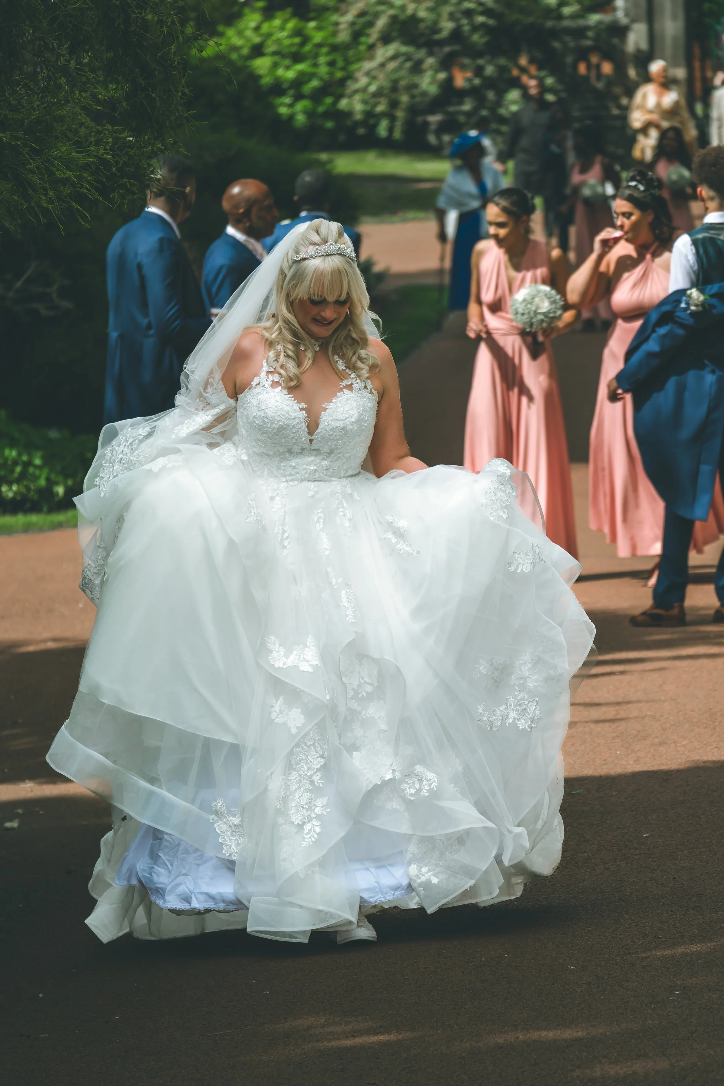 A bride in a white wedding gown is holding up her dress while walking outdoors on a paved path, with bridesmaids and groomsmen standing in the background.