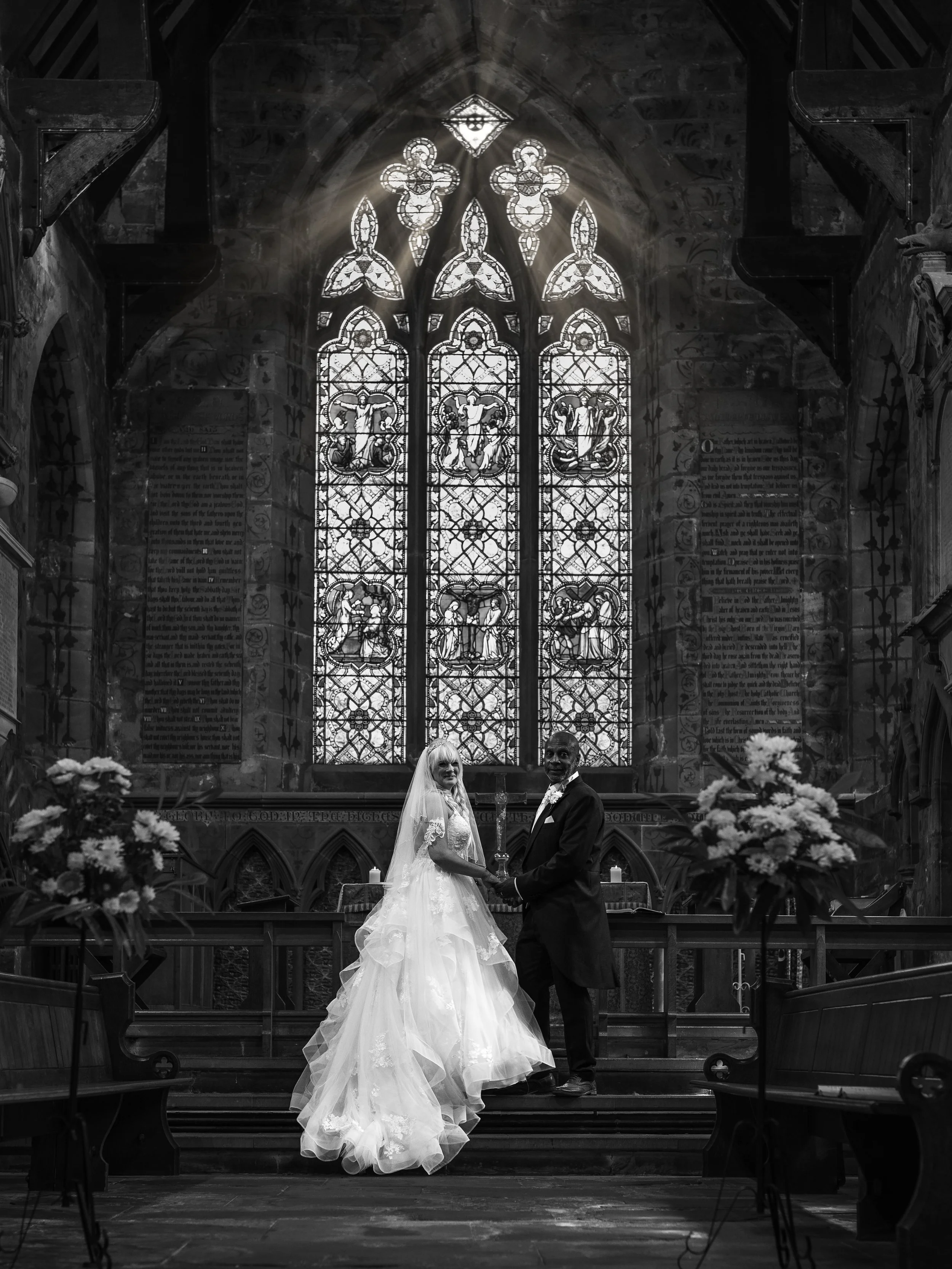 A bride and groom standing hand in hand inside a church, in front of a large stained-glass window, surrounded by flowers and lit candles.
