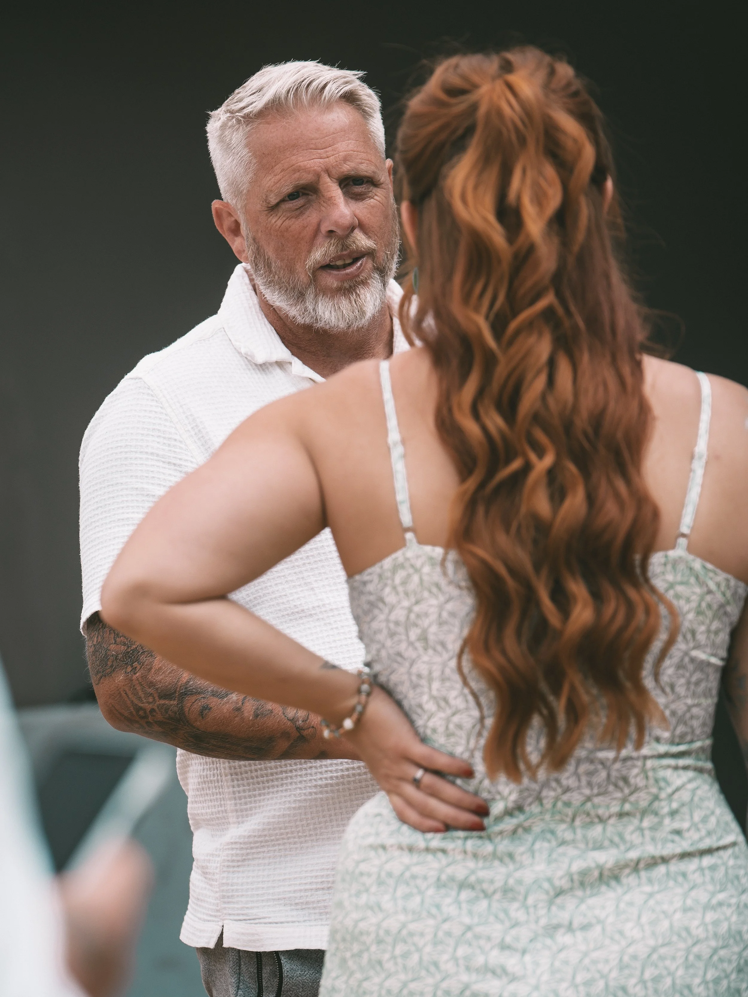 An older man with gray hair and a beard, wearing a white shirt, engaging in a conversation with a woman with long red curly hair wearing a patterned dress. They are outdoors.