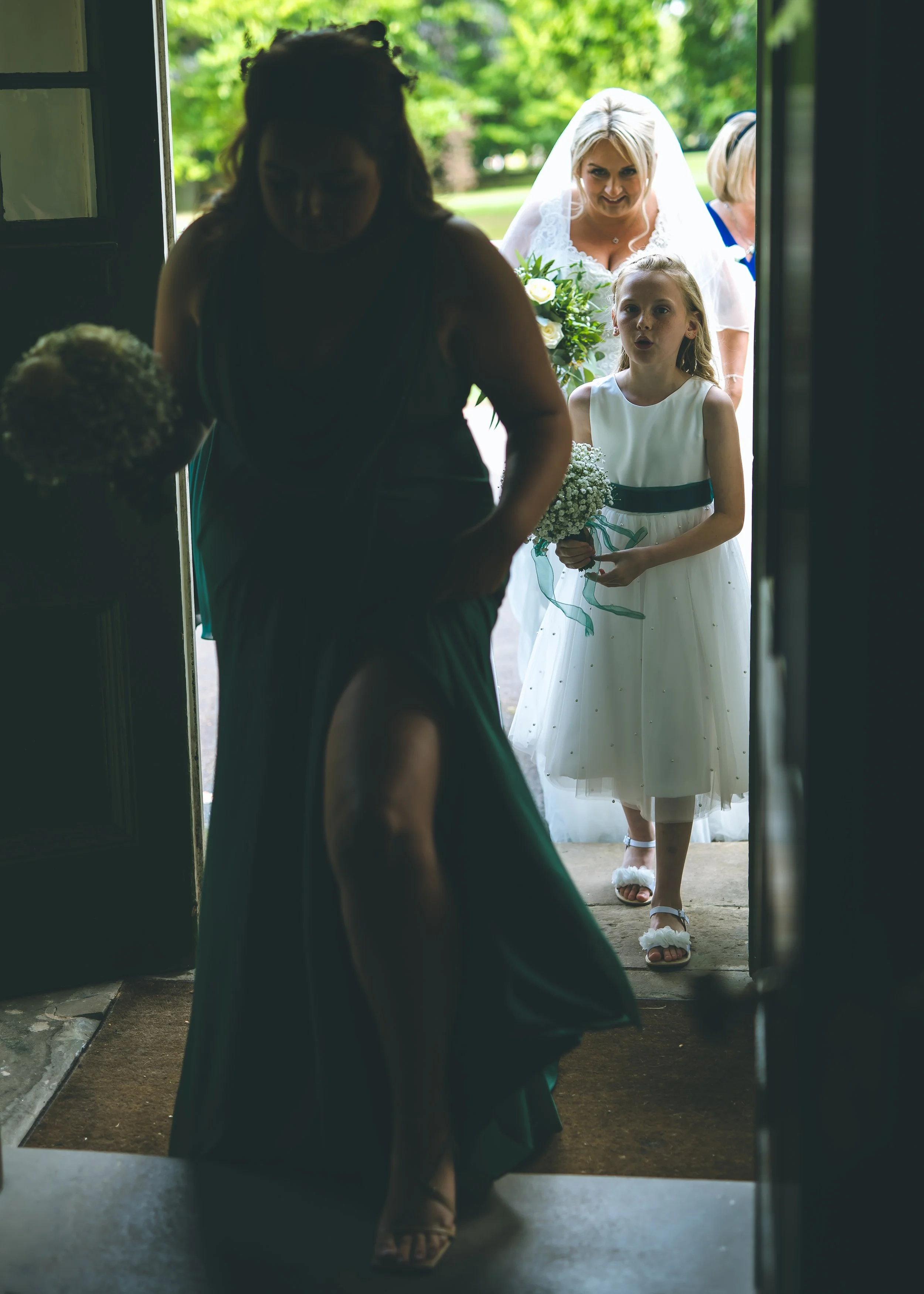 A bride in a white wedding dress and veil is arriving at a wedding venue, holding a bouquet, as she enters through a doorway. A young girl in a white dress with dark ribbon, holding a small bouquet, is standing in the doorway, with other women and lu
