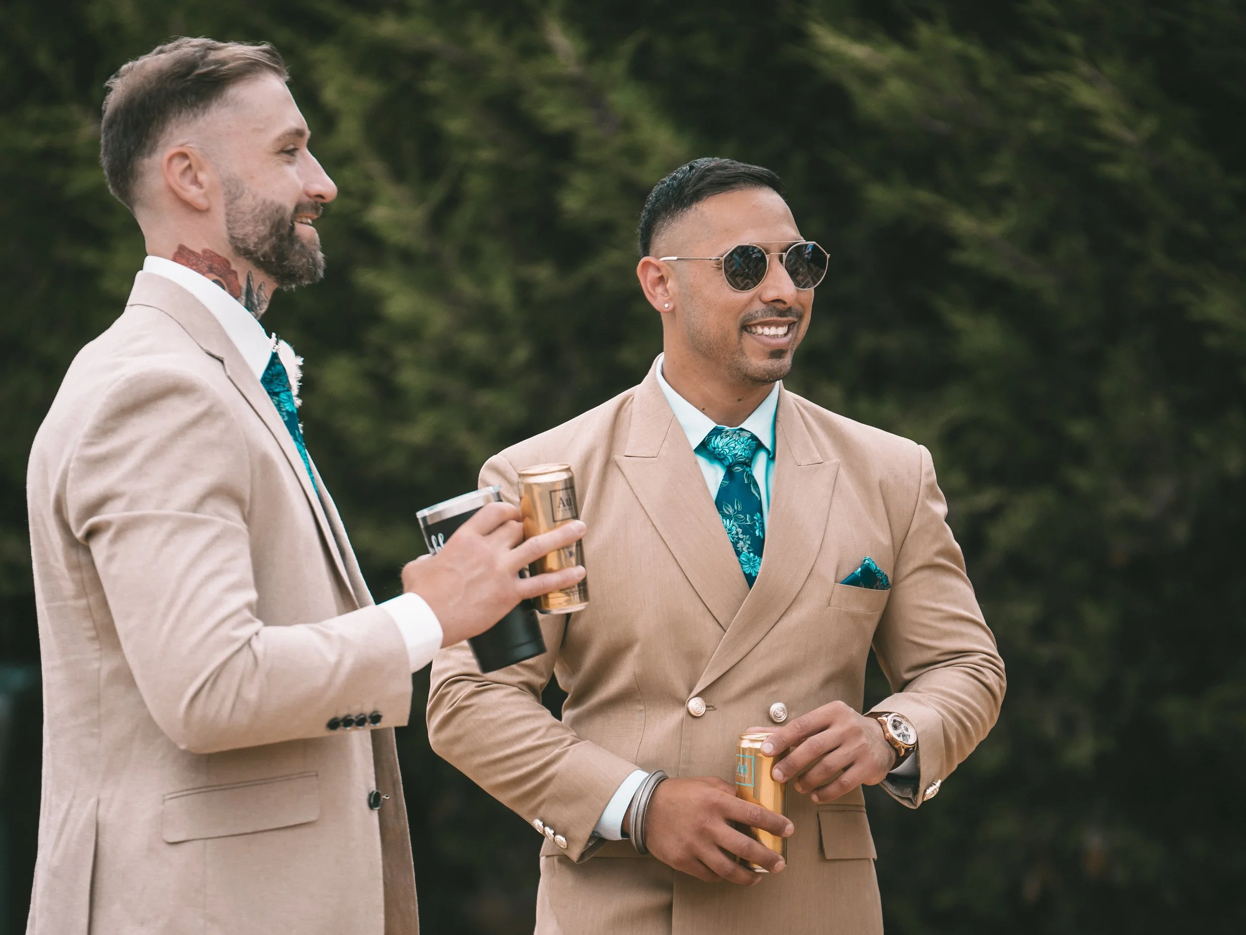 Two men in beige suits stand outdoors, smiling and engaging in conversation, each holding a drink. One man has short dark hair, sunglasses, and a blue patterned tie, while the other has a beard, tattoos, and light brown hair.