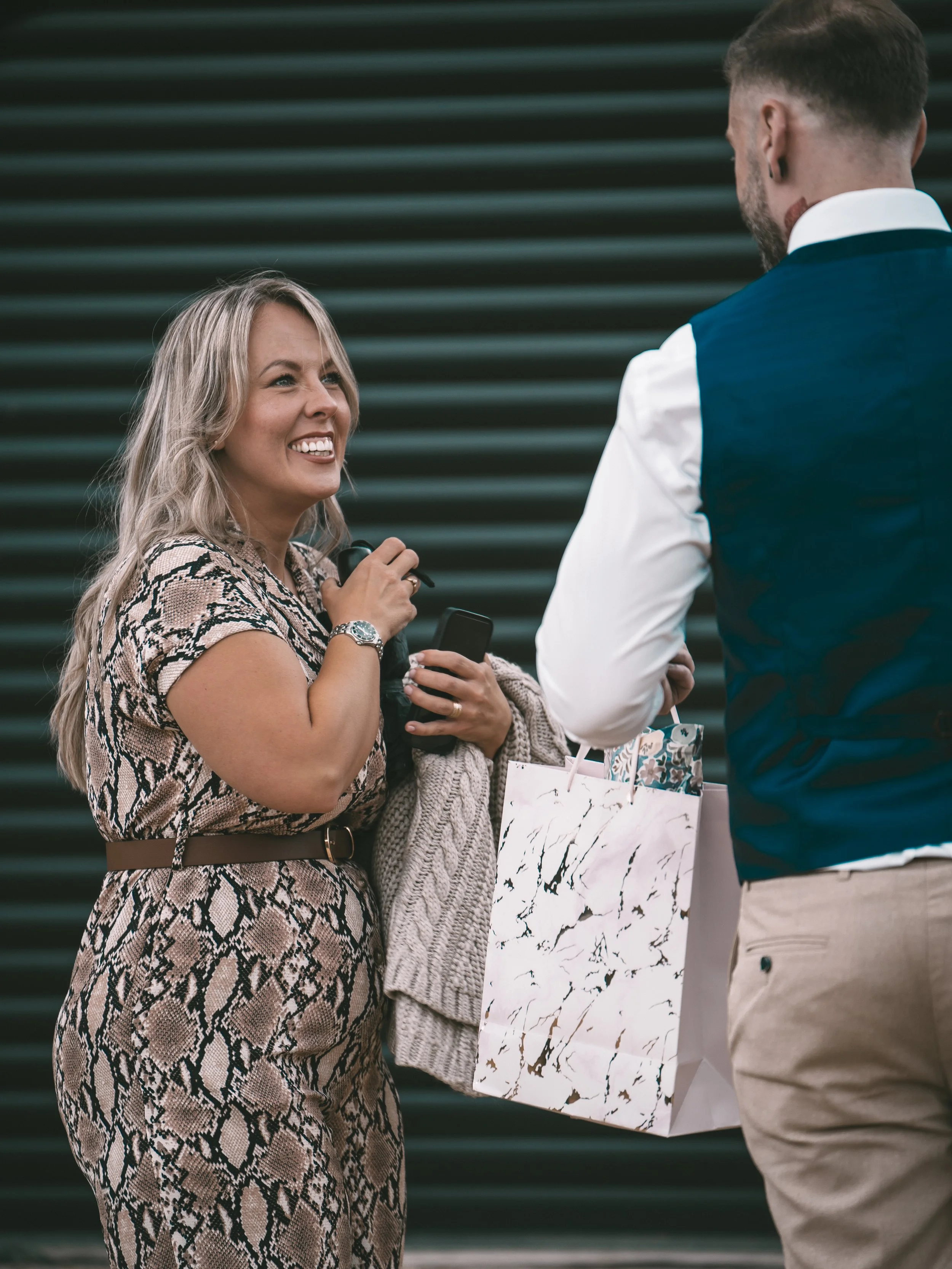 A woman with blonde hair smiling and holding a microphone stands next to a man in a blue vest and cream-colored pants, who is holding a gift bag. They are engaged in conversation in front of a dark striped background.