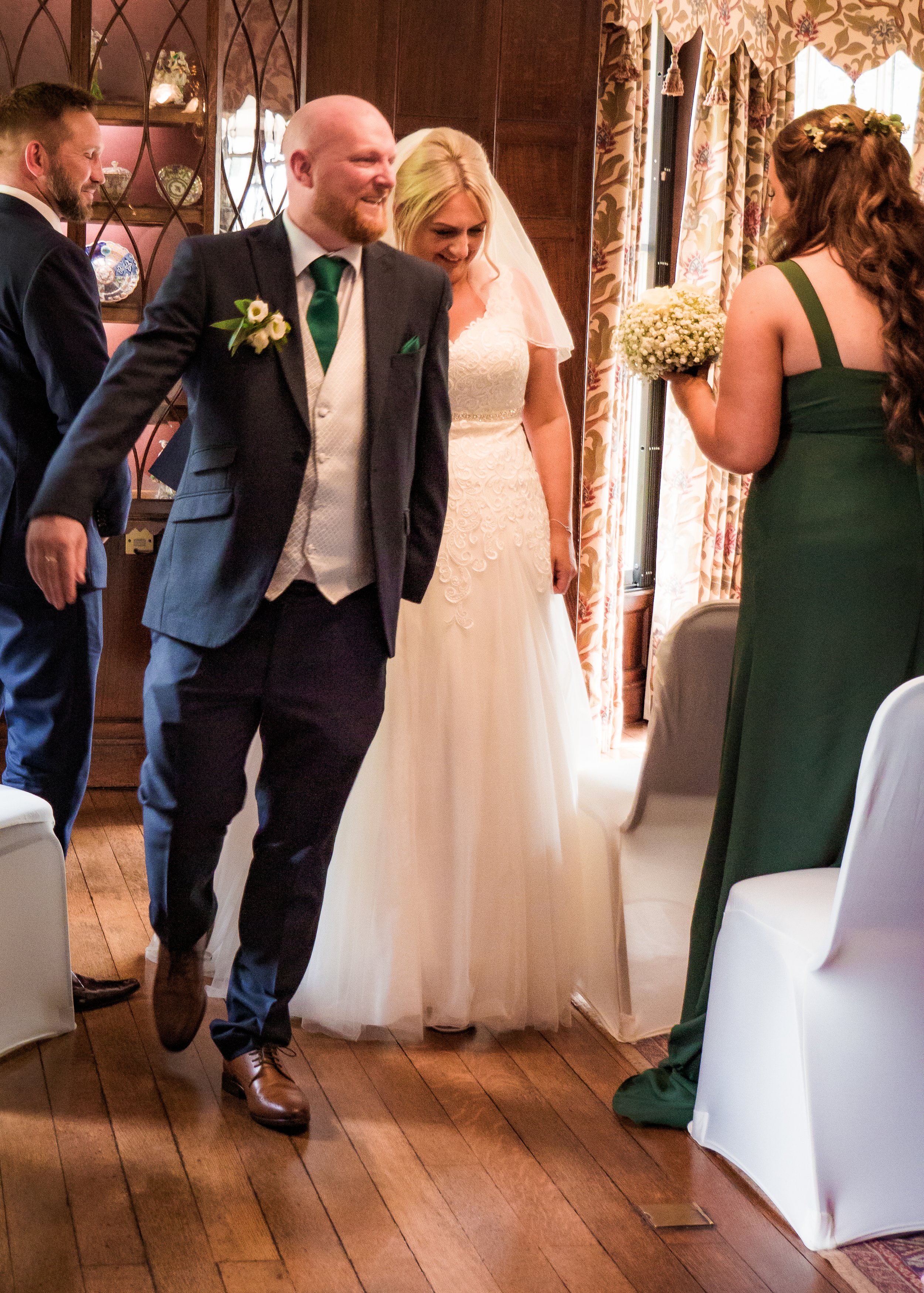 A bride in a white wedding dress with angel wings looks while a woman in a green dress holds a bouquet, and two men in suits are nearby, inside a decorated room with wooden walls and patterned curtains.