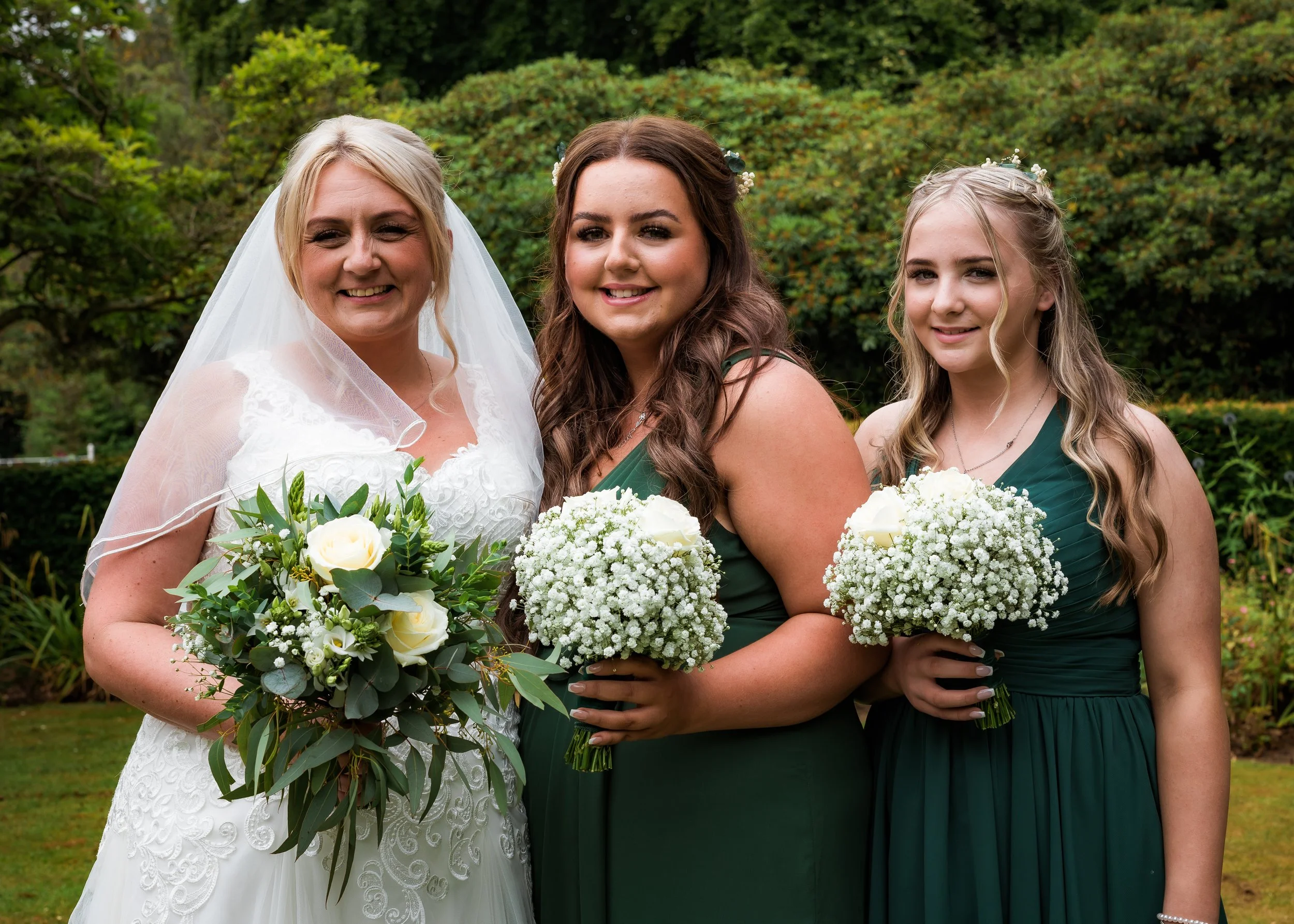 A bride standing with two women outdoors, holding bouquets of white flowers, smiling in front of green foliage.