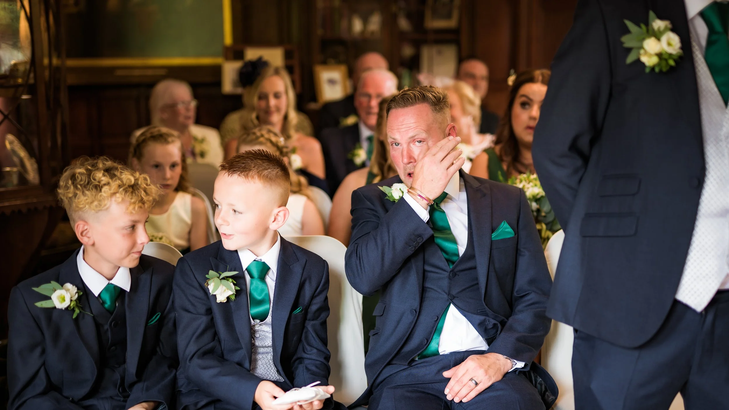 Group of people sitting at a wedding ceremony, two young boys and a man in formal attire with boutonnieres, sitting with other guests in the background.