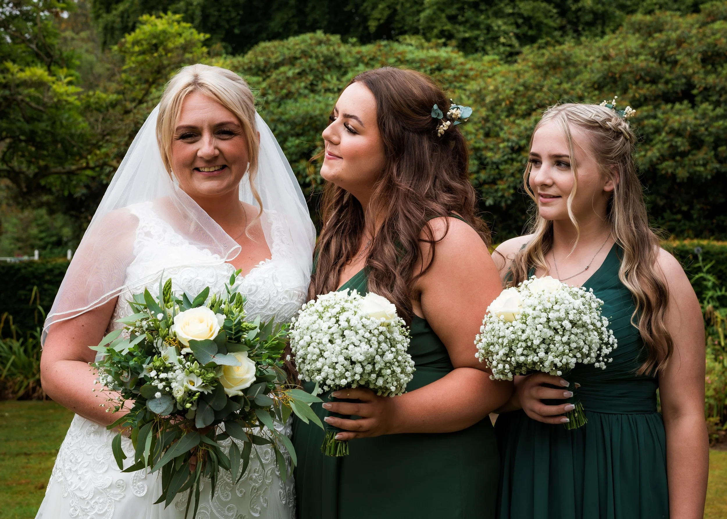 A bride and two bridesmaids holding bouquets of white flowers and greenery outdoors in front of green bushes.