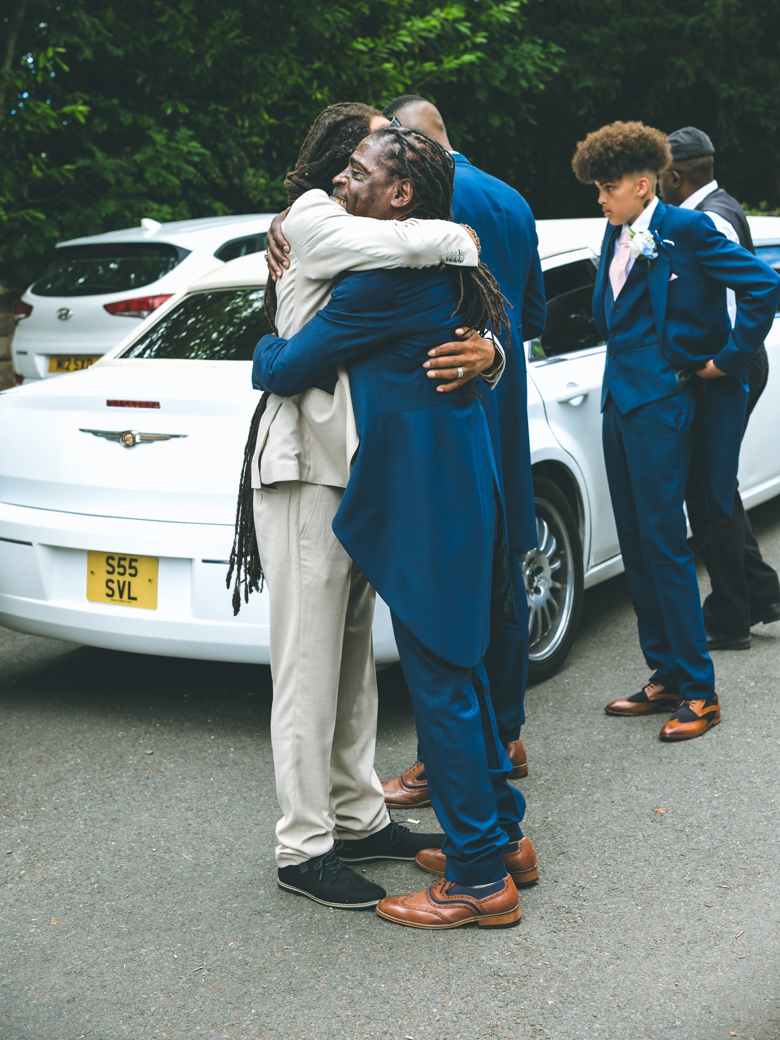 Two men hugging in front of a white car, with two other men standing nearby, one younger and one older, dressed in formal suits at an outdoor event.