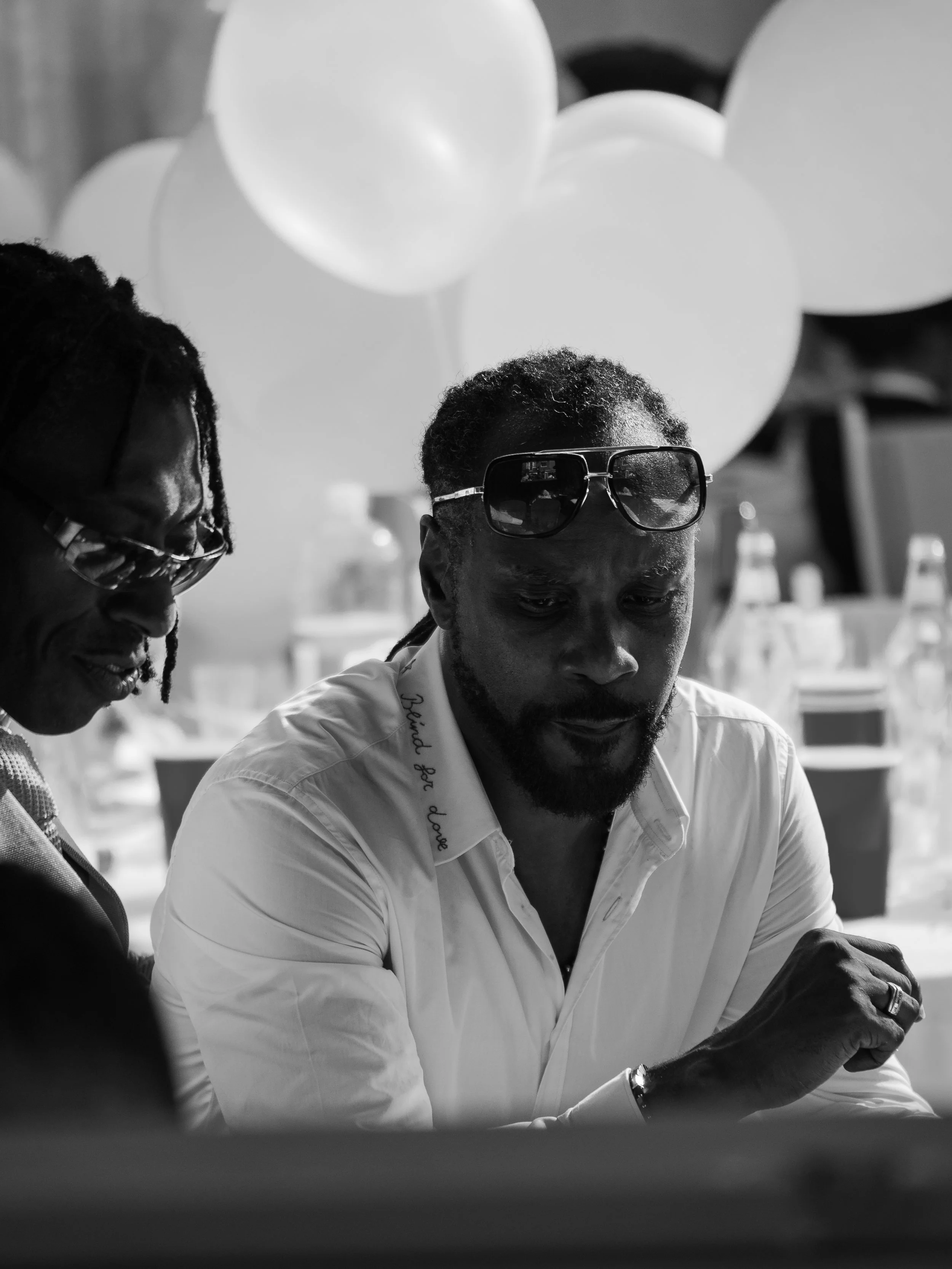 Two men are engaged in an activity, with one wearing sunglasses and a white shirt and the other with glasses and dreadlocks, in a room with balloons and bottles in the background.