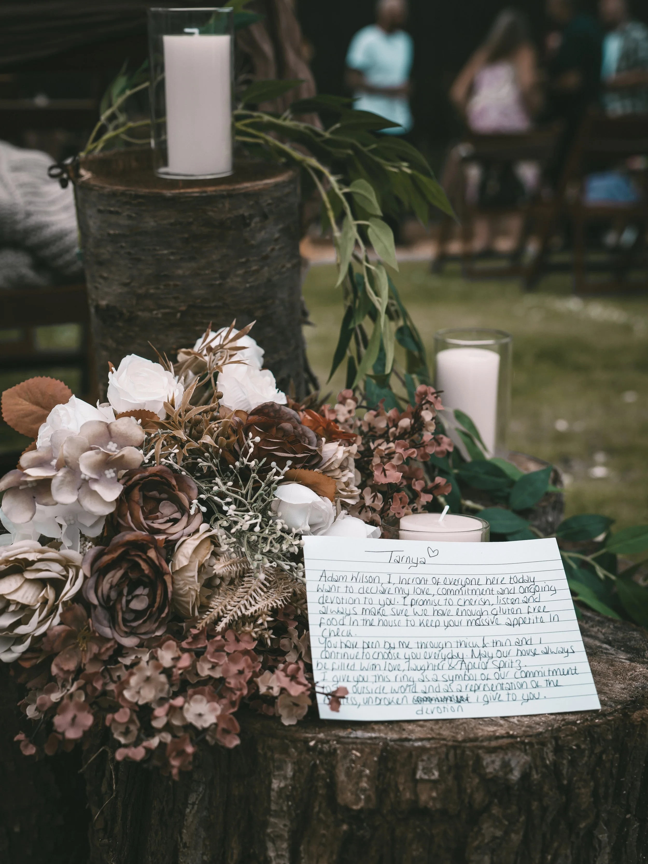 A floral arrangement with white and brown flowers and greenery on a wooden stump, with a handwritten letter and candles nearby, at an outdoor event.
