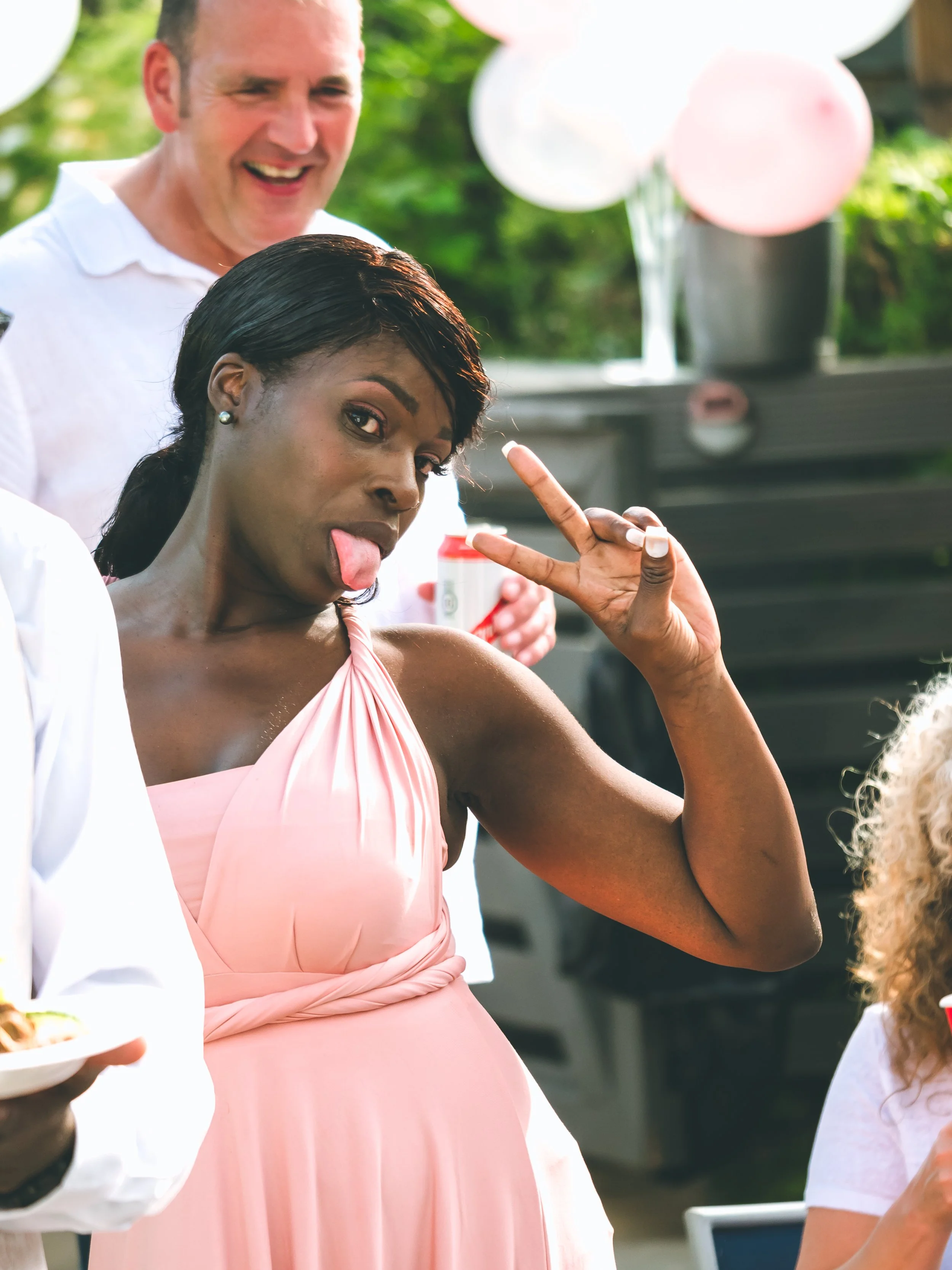 Woman in pink dress making a peace sign with her hand at an outdoor gathering with balloons and people around.