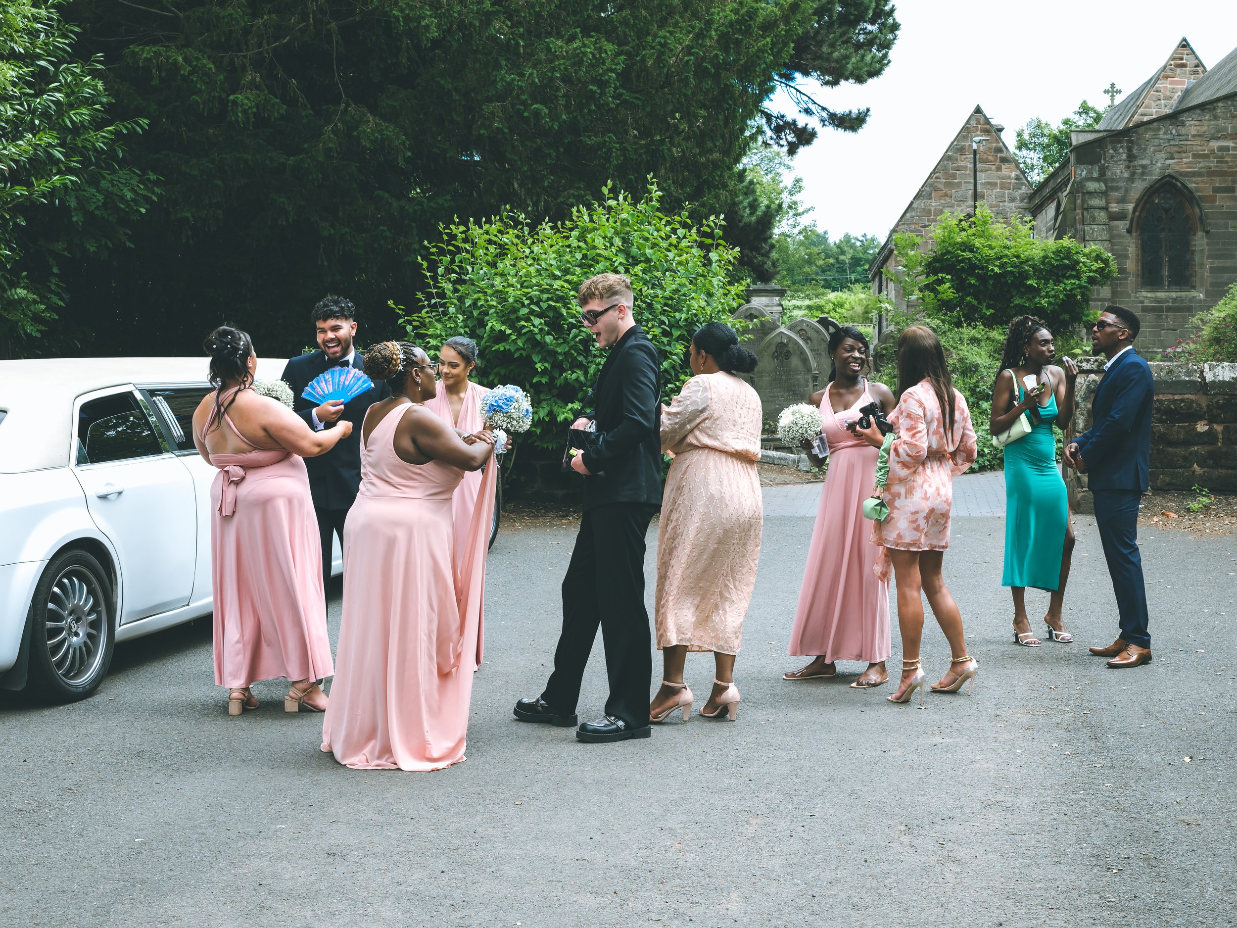 A diverse group of people dressed in pastel-colored dresses and suits are gathered outdoors near a church with stone walls and a graveyard. They are socializing, taking photos, and exchanging greetings, indicating a wedding celebration.
