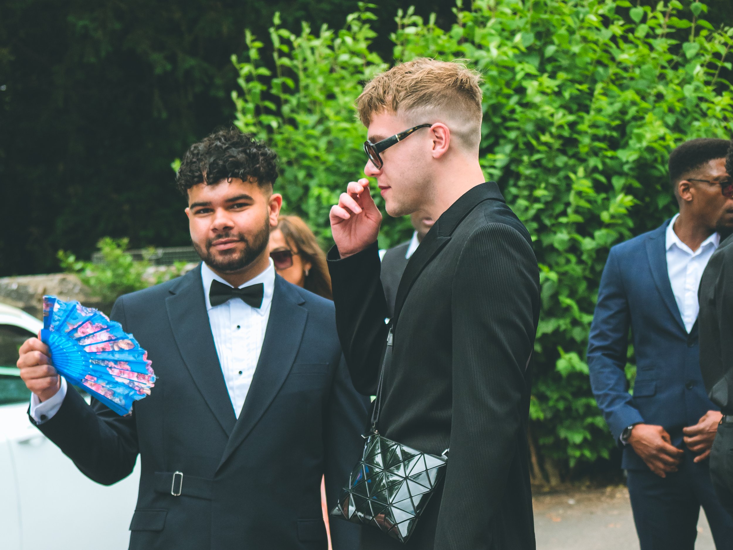 Two men in formal suits standing outdoors. The man on the left has dark curly hair, a beard, and is holding a colorful fan. The man on the right has light brown hair, glasses, and is adjusting his glasses. There are other people and greenery in the b
