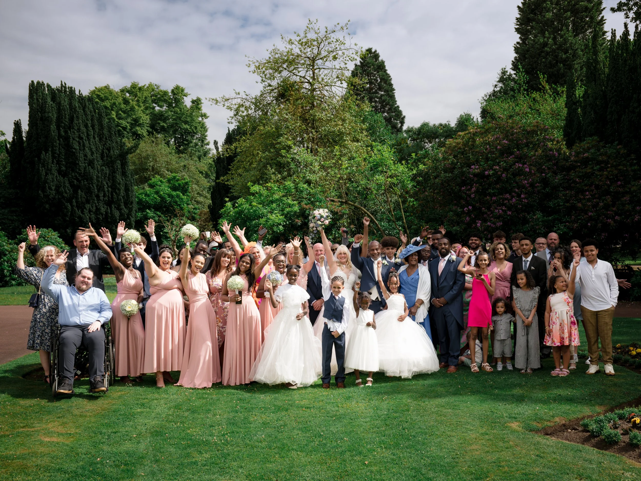 A large diverse group of people celebrating outdoors in a park, with many raising their hands, some holding bouquets, with trees and a cloudy sky in the background.