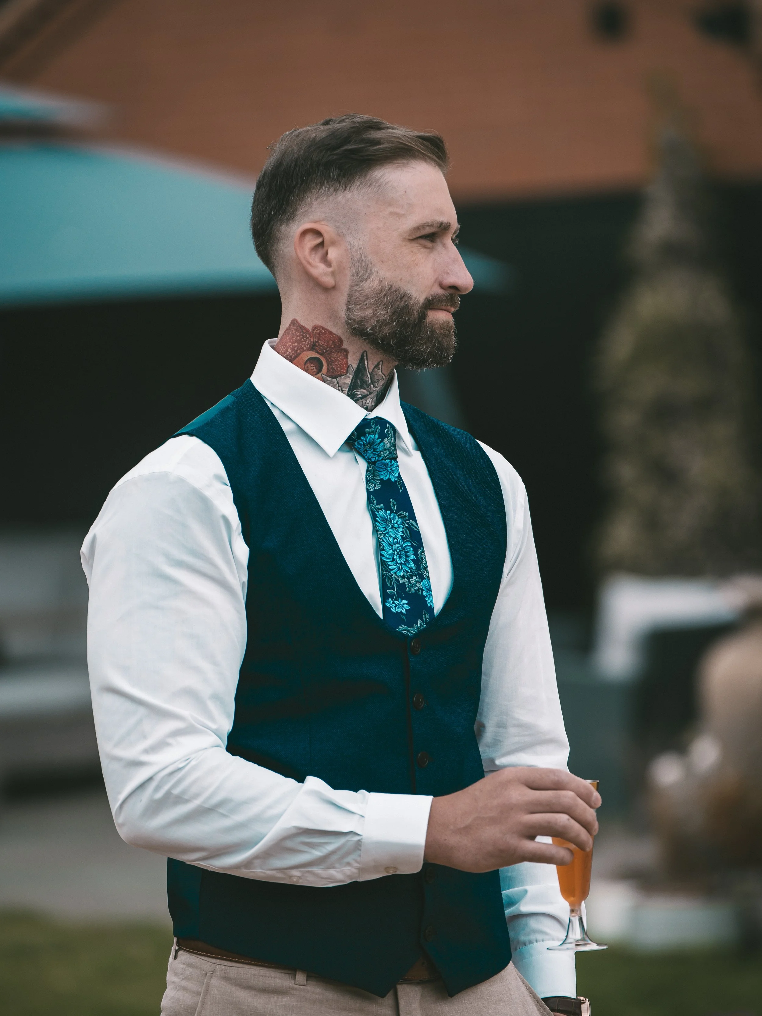 A man with a beard and tattoos on his neck, dressed in a white shirt and a dark vest, holding a drink in a glass with a straw, at an outdoor event.