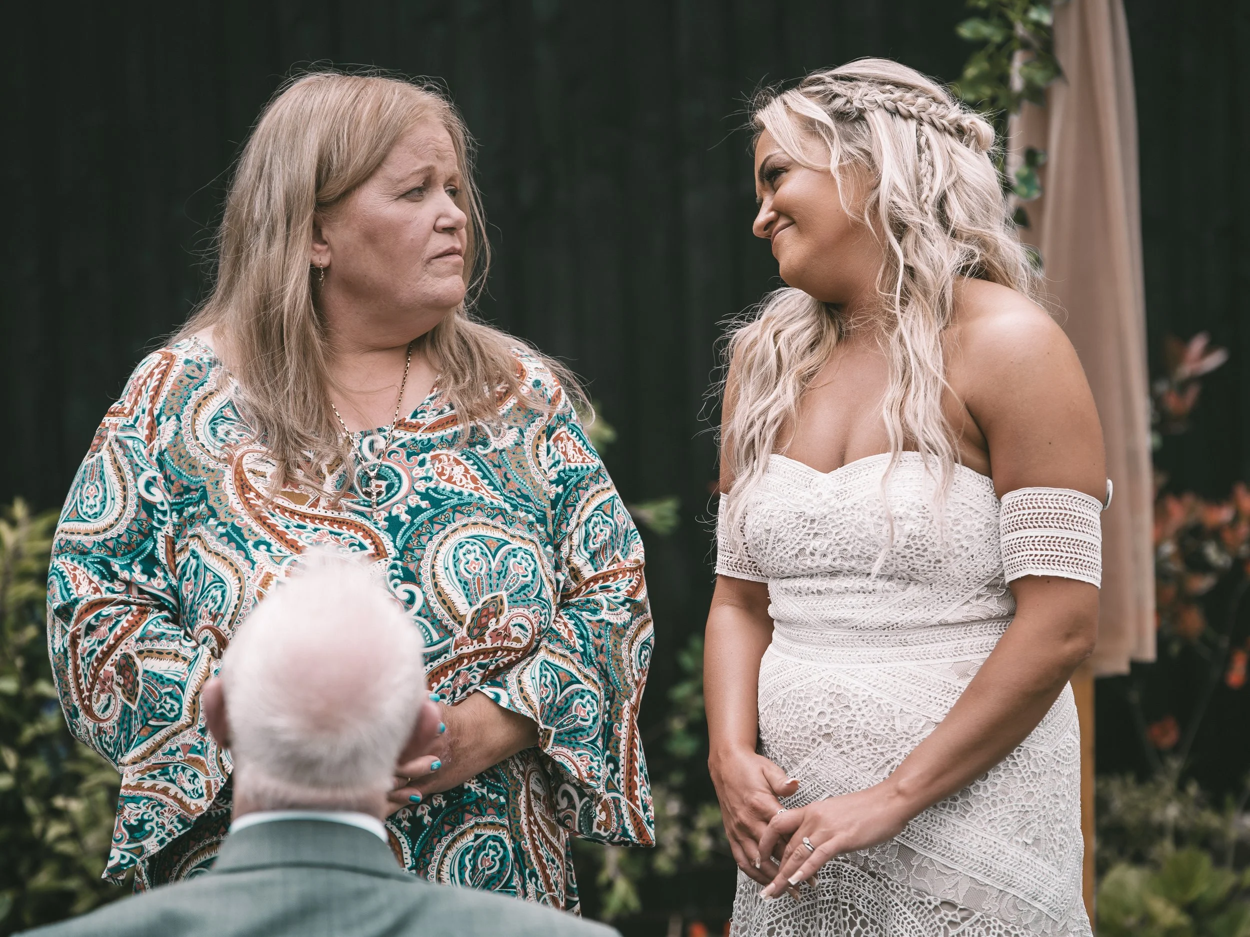A woman with light blonde hair, braided on one side, in a white lace dress standing with her hands clasped, smiling at another woman who has light brown hair, wearing a colorful paisley blouse with a necklace, during a wedding ceremony outdoors.