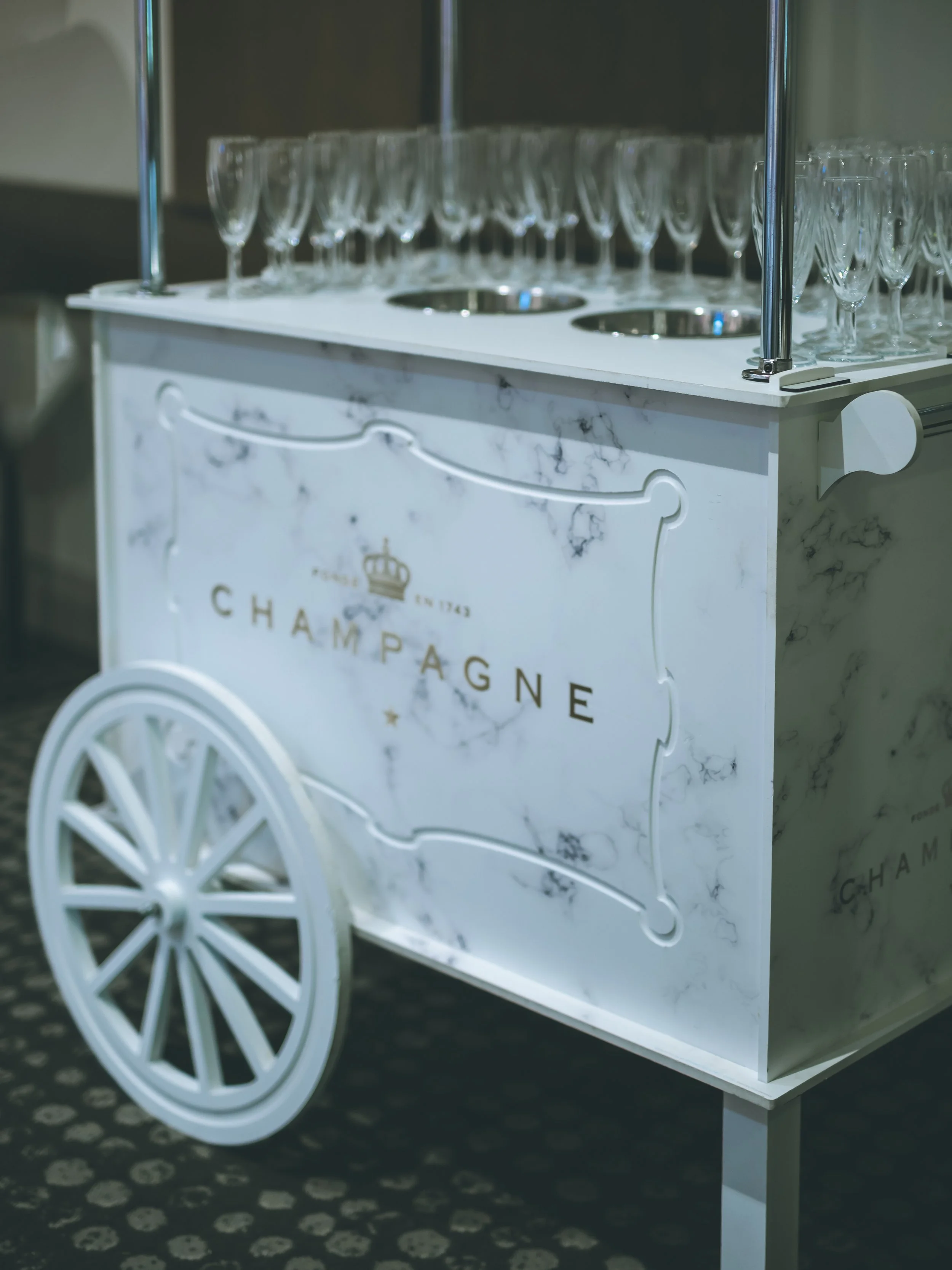 A white marble-textured bar cart with the words 'Champagne' and a crown printed on it, featuring large white wheels, and filled with empty champagne glasses, set on a black and white patterned carpet.