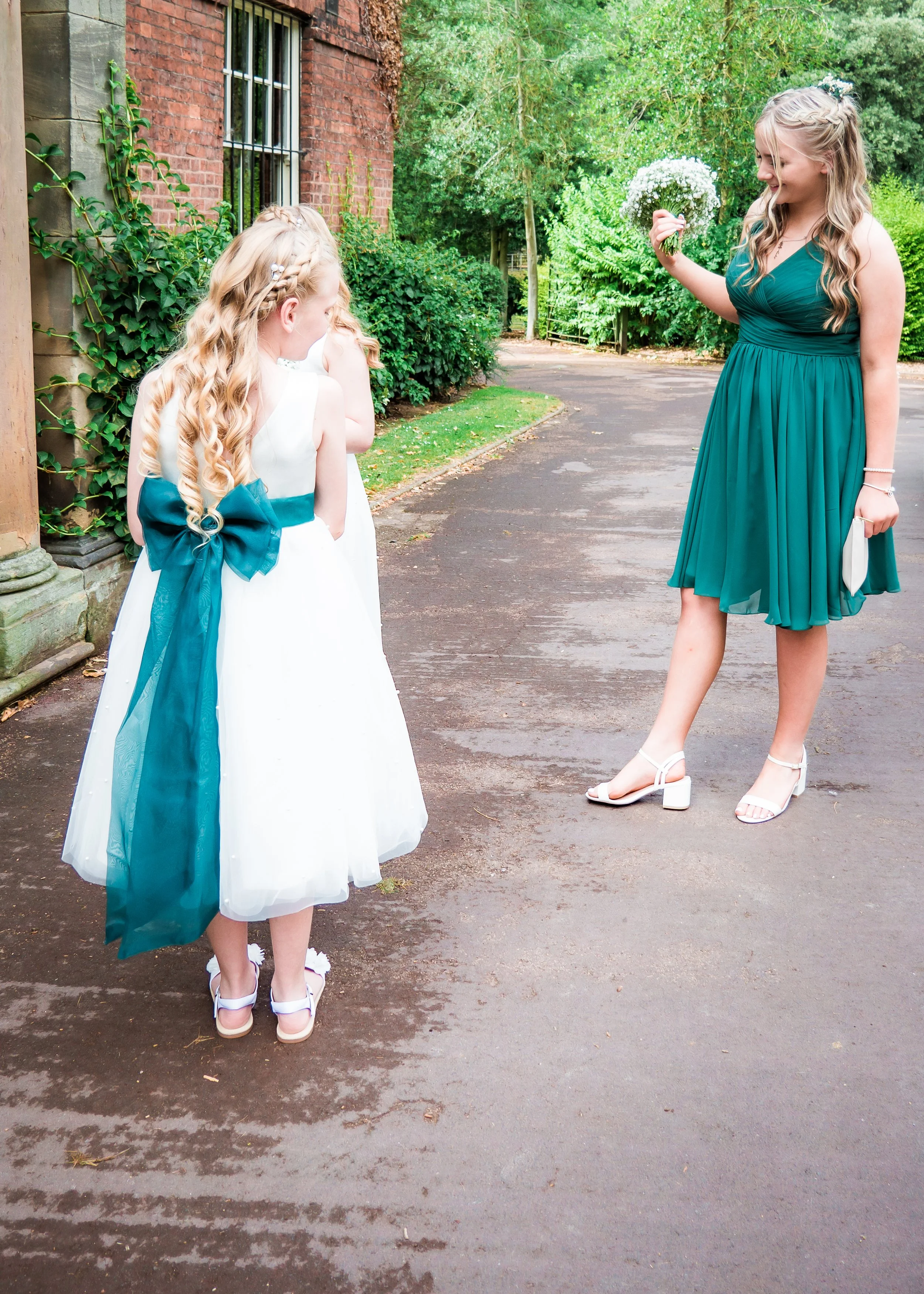 A woman in a teal dress giving a bouquet of white flowers to two young girls dressed in white dresses with teal bows, outside near a brick building and green trees.