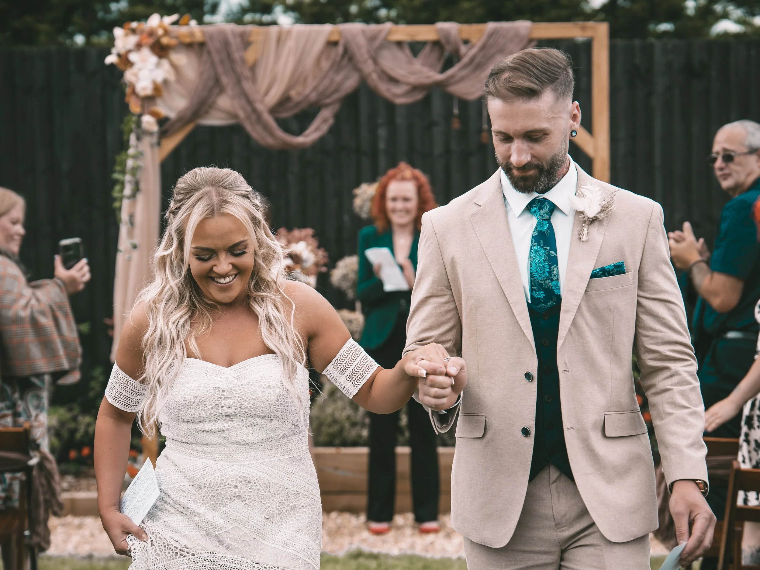 A couple getting married outdoors, holding hands with wedding rings visible, standing in front of a ceremony arch decorated with fabric and flowers, surrounded by guests.