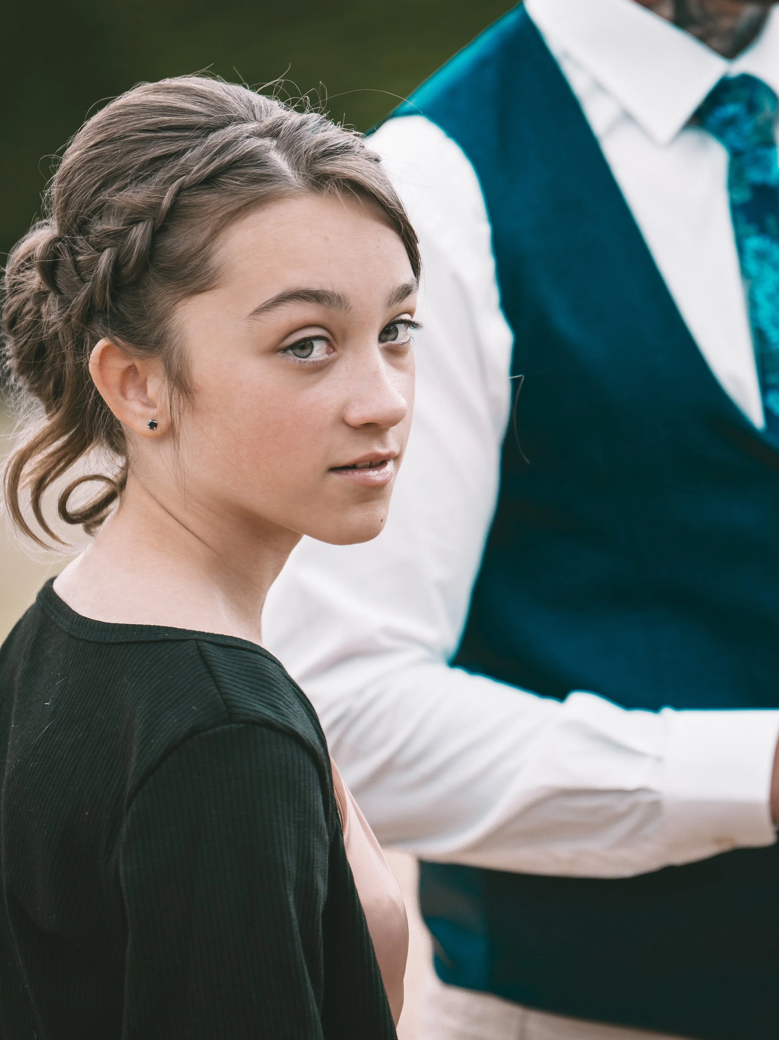 Close-up of a young girl with braided hair and earrings, looking slightly to the side, wearing a black top with another person partially visible beside her, wearing a white shirt and blue vest.