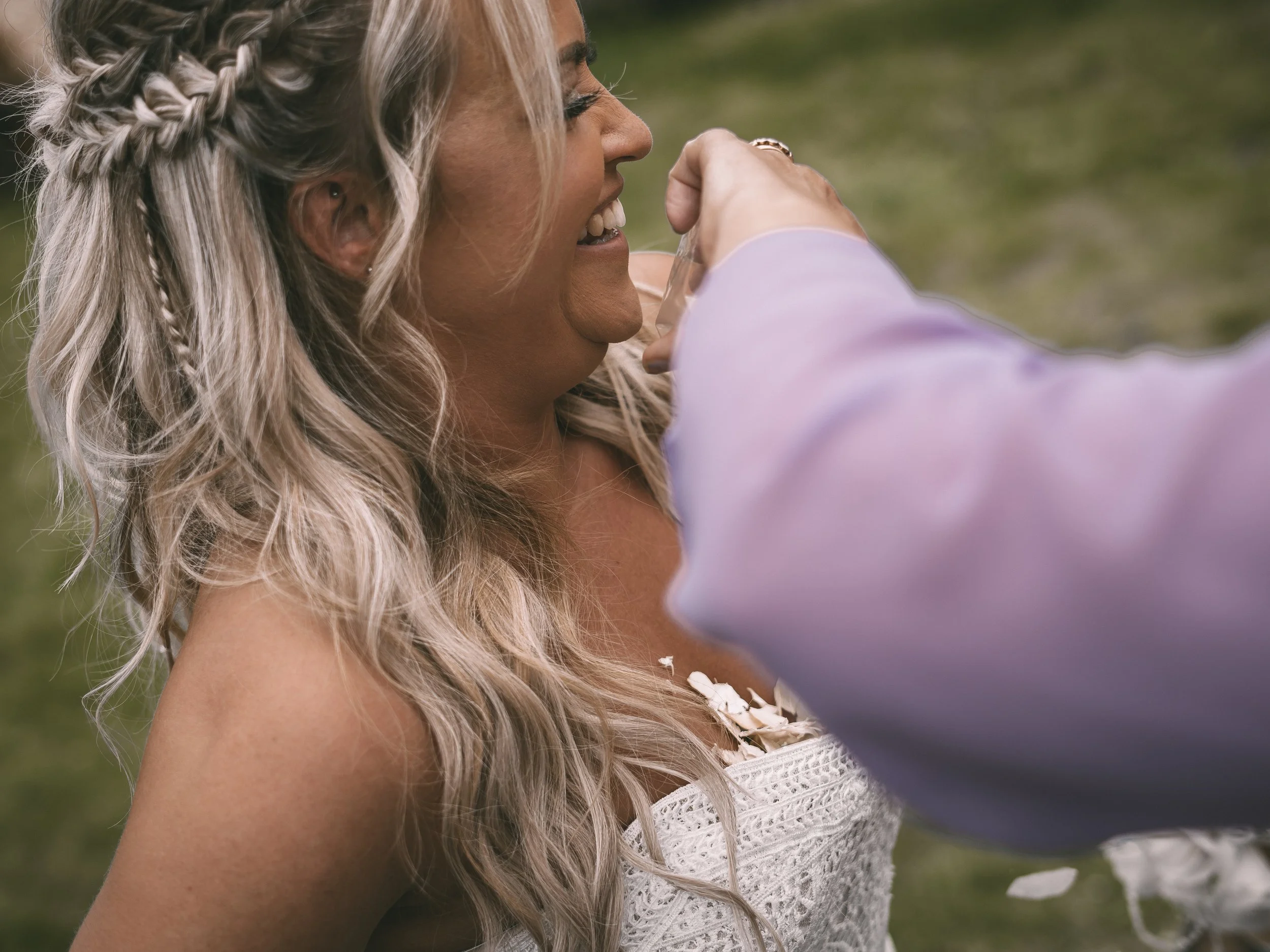 A woman with long, wavy, blonde hair styled in braids smiling and smiling with a person in a lavender shirt touching her face outdoors near green grass.