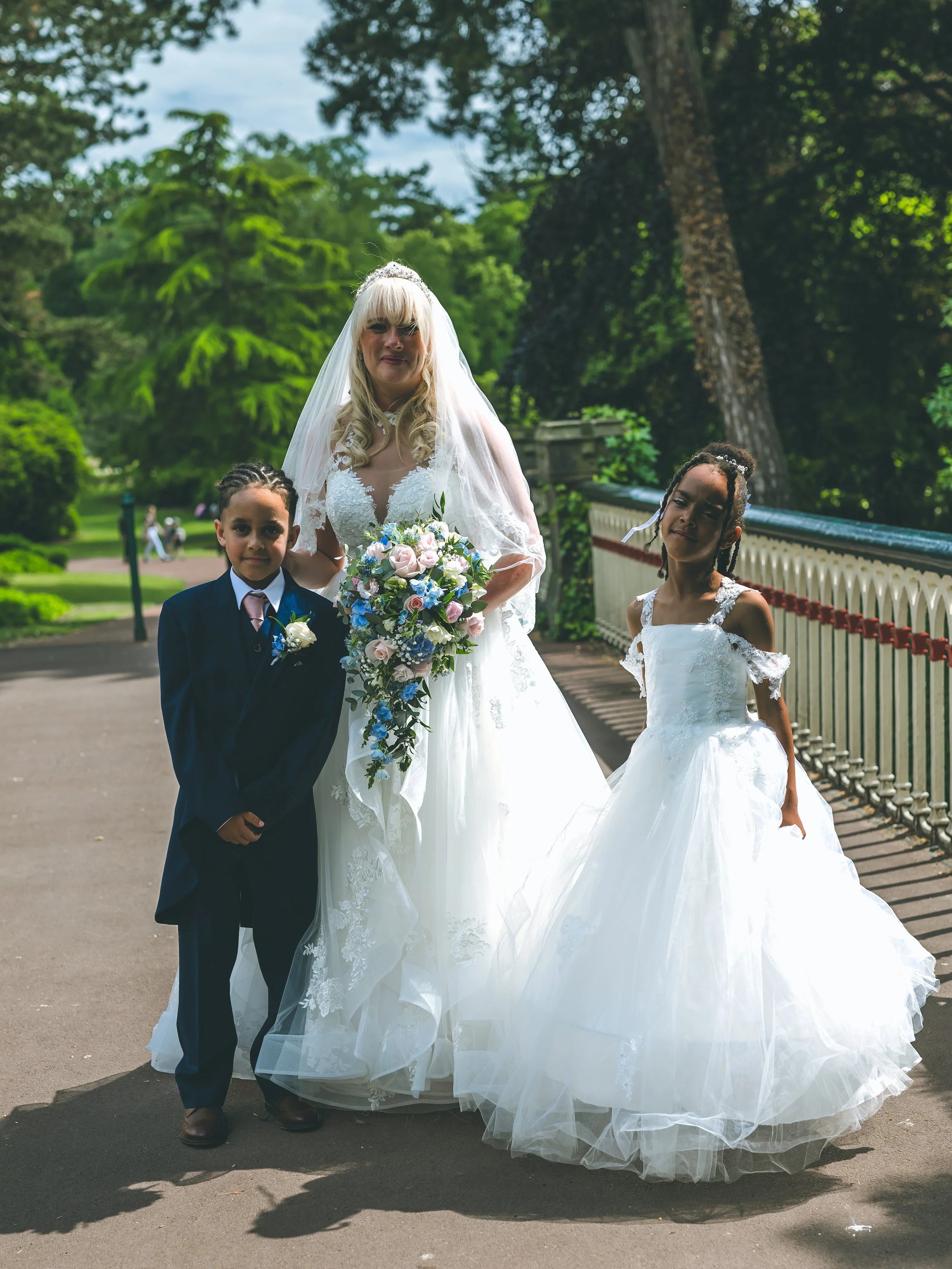 A woman in a wedding dress holding a bouquet of flowers, standing outdoors with two young girls, all dressed in formal attire, on a bridge with trees in the background.