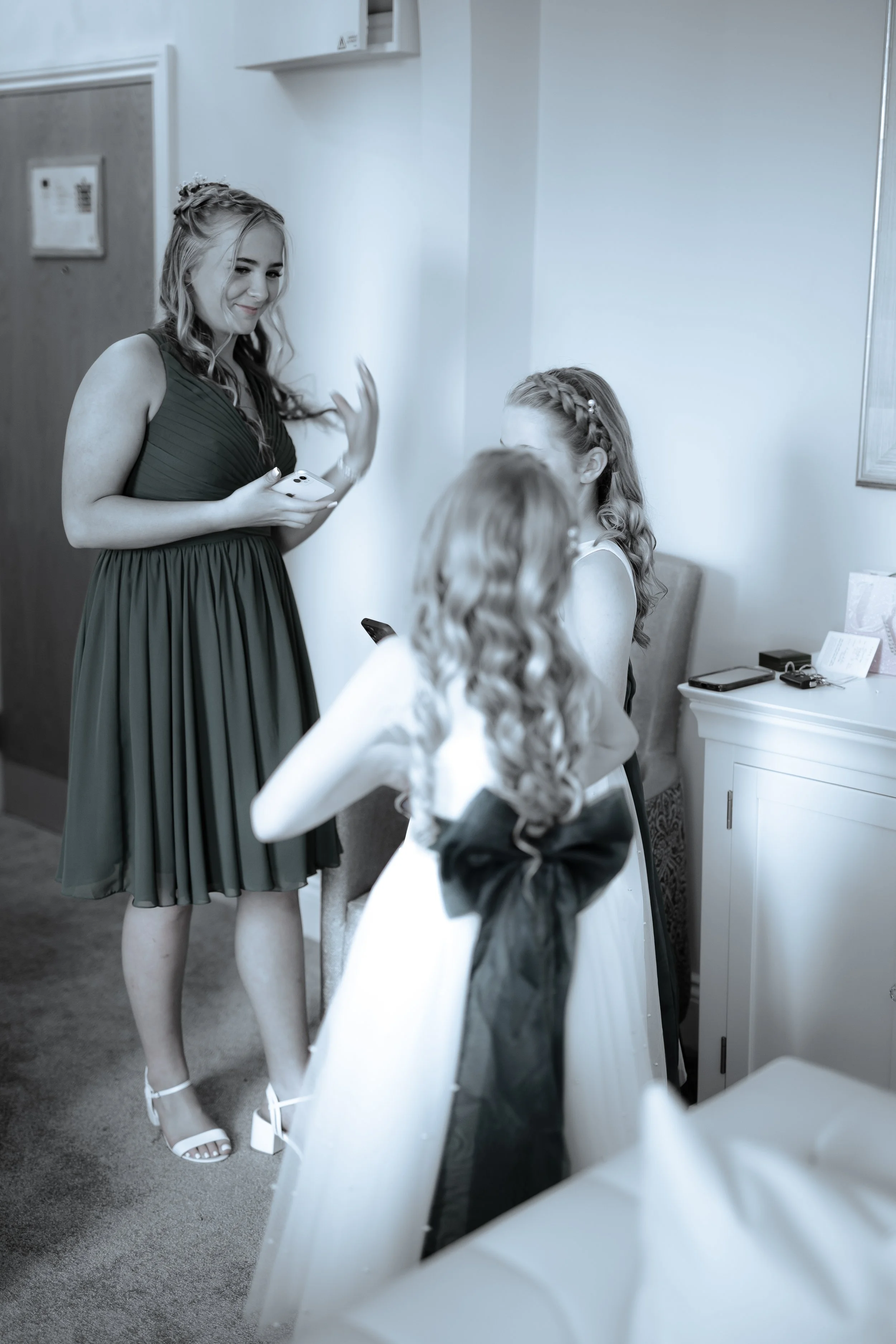 A woman in a dark green dress with white heels is smiling and gesturing with her hand while talking to two other women in white dresses with braided hair, seated near a white table in an indoor setting.