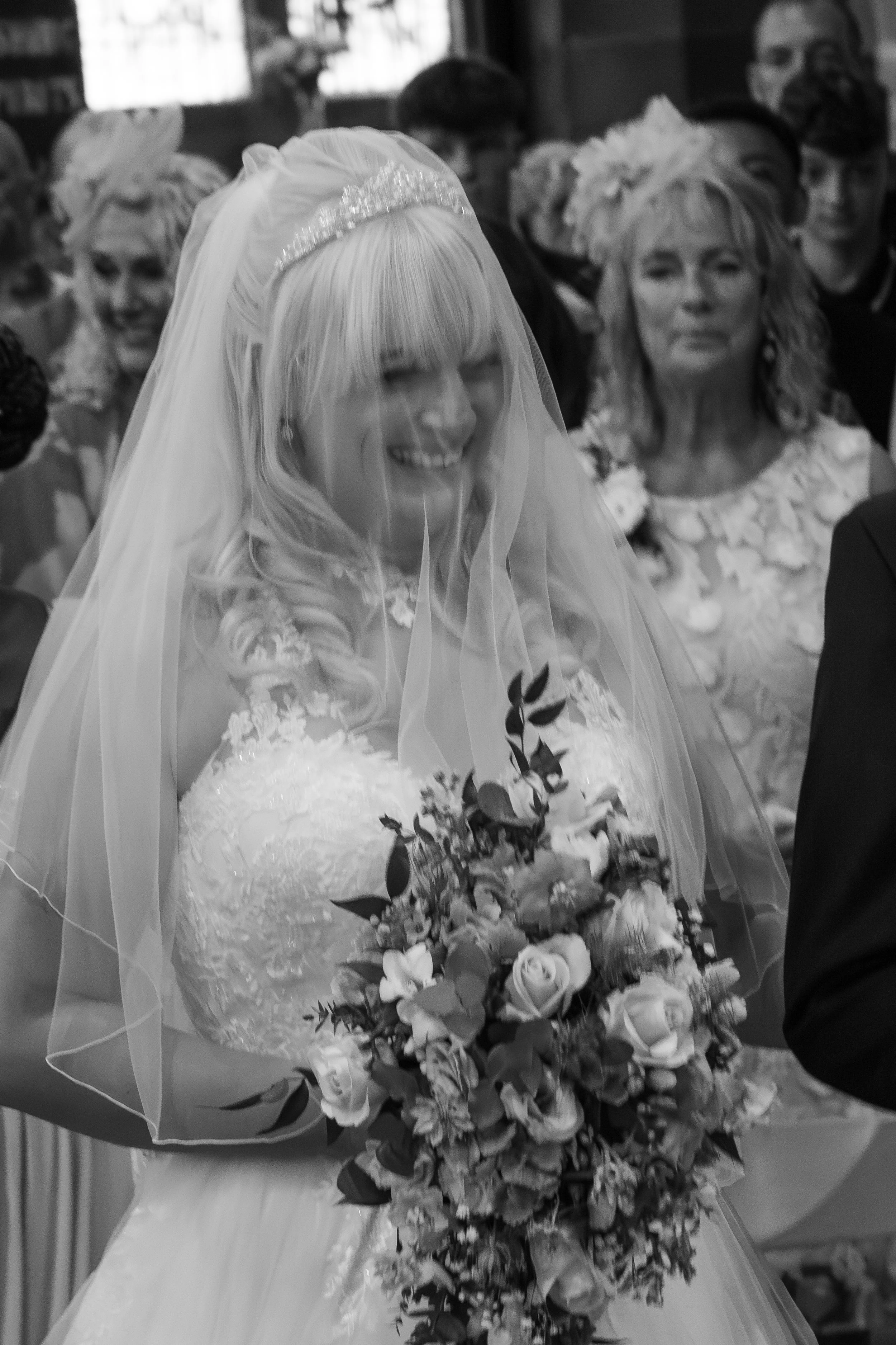 A bride with light blonde hair wearing a veil, wedding dress, and tiara, smiling and holding a large bouquet of roses and greenery. Behind her are guests, including women with floral headpieces, in a celebration setting.