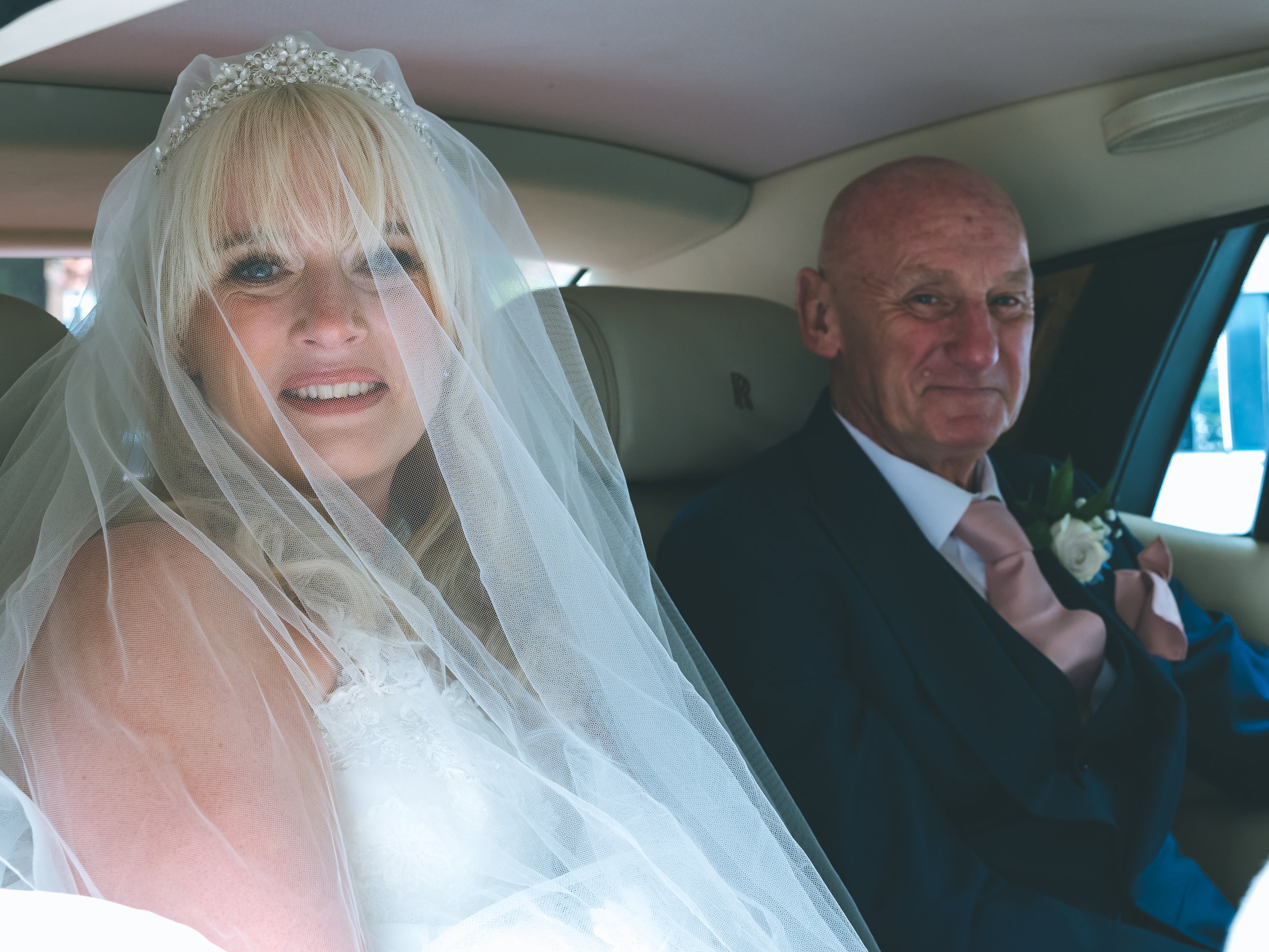 A bride with blonde hair and a lace veil smiling inside a vehicle, seated next to a groom in a suit holding a boutonniere, both dressed for a wedding.