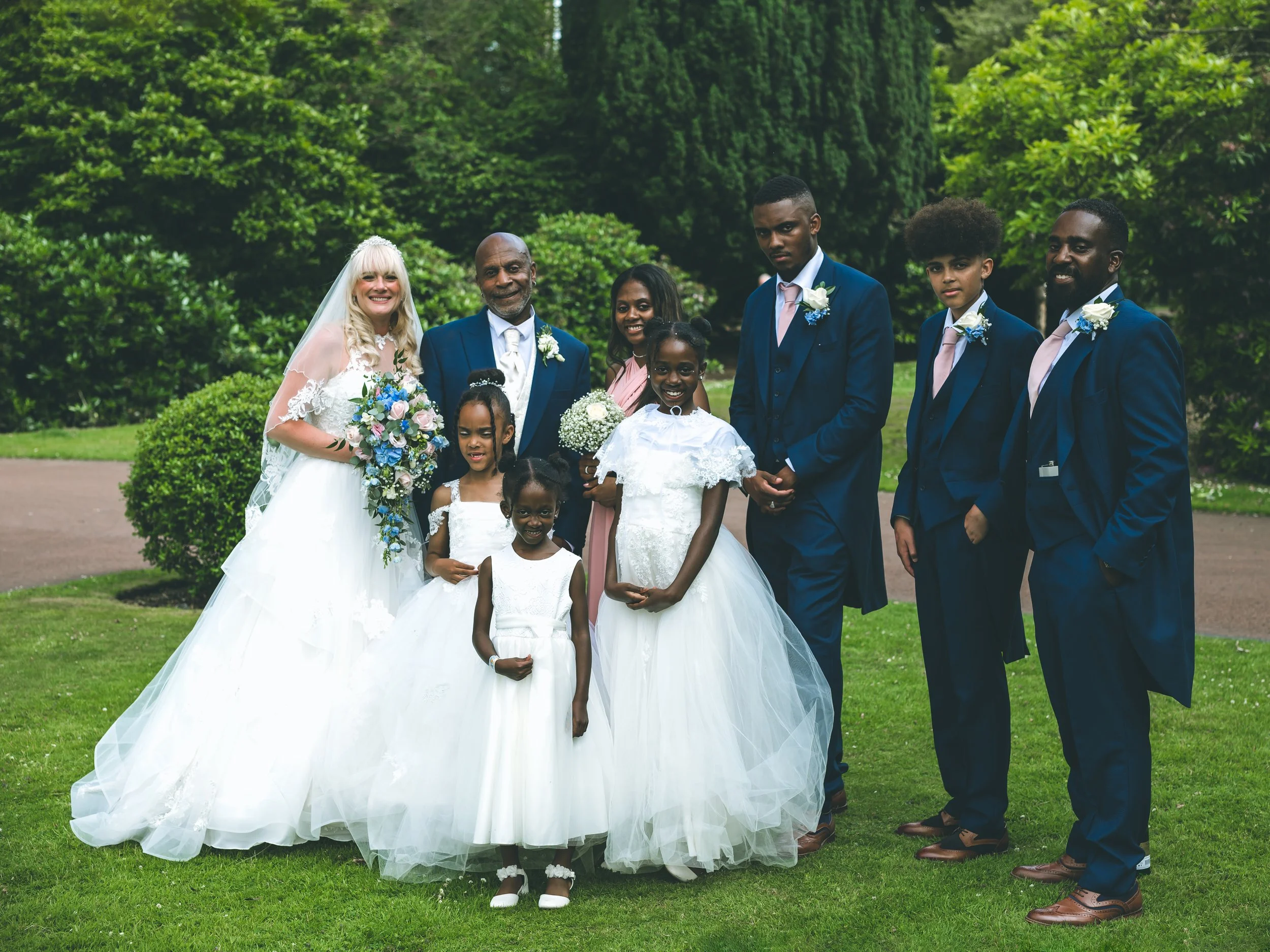 A group of people dressed for a wedding, including a bride in a white gown and veil holding a bouquet, standing outdoors on a grassy area surrounded by green trees.