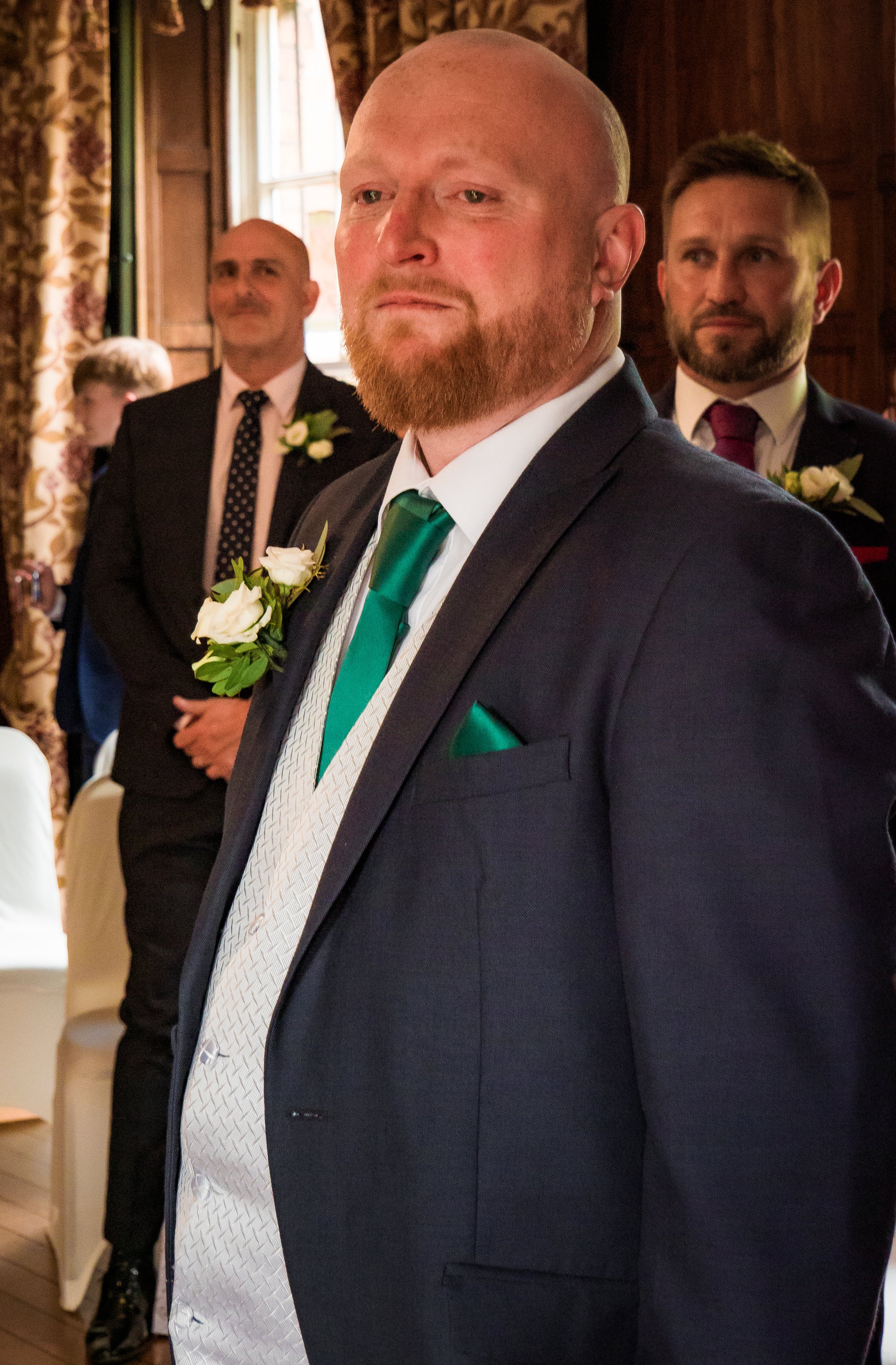 A group of men in formal suits, likely at a wedding or similar event, with one man in the foreground wearing a dark suit, white shirt, and green tie, and others in the background wearing suits and boutonnières, inside a wood-paneled room with a windo