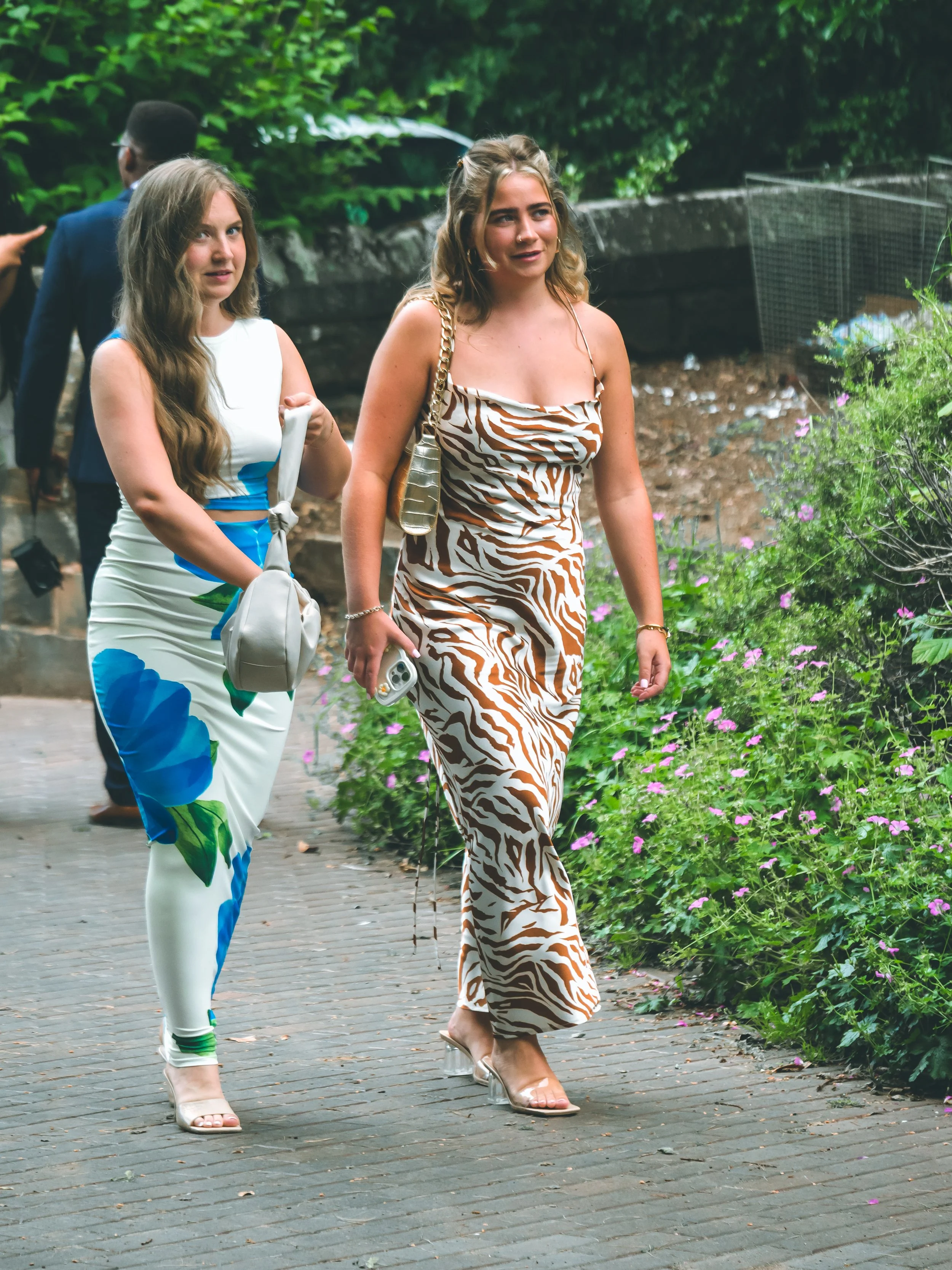 Two women walking along a sidewalk with greenery and flowers. One woman is wearing a white dress with blue and green abstract patterns, and the other is wearing a tiger print dress with clear heeled shoes. Both women are carrying handbags.