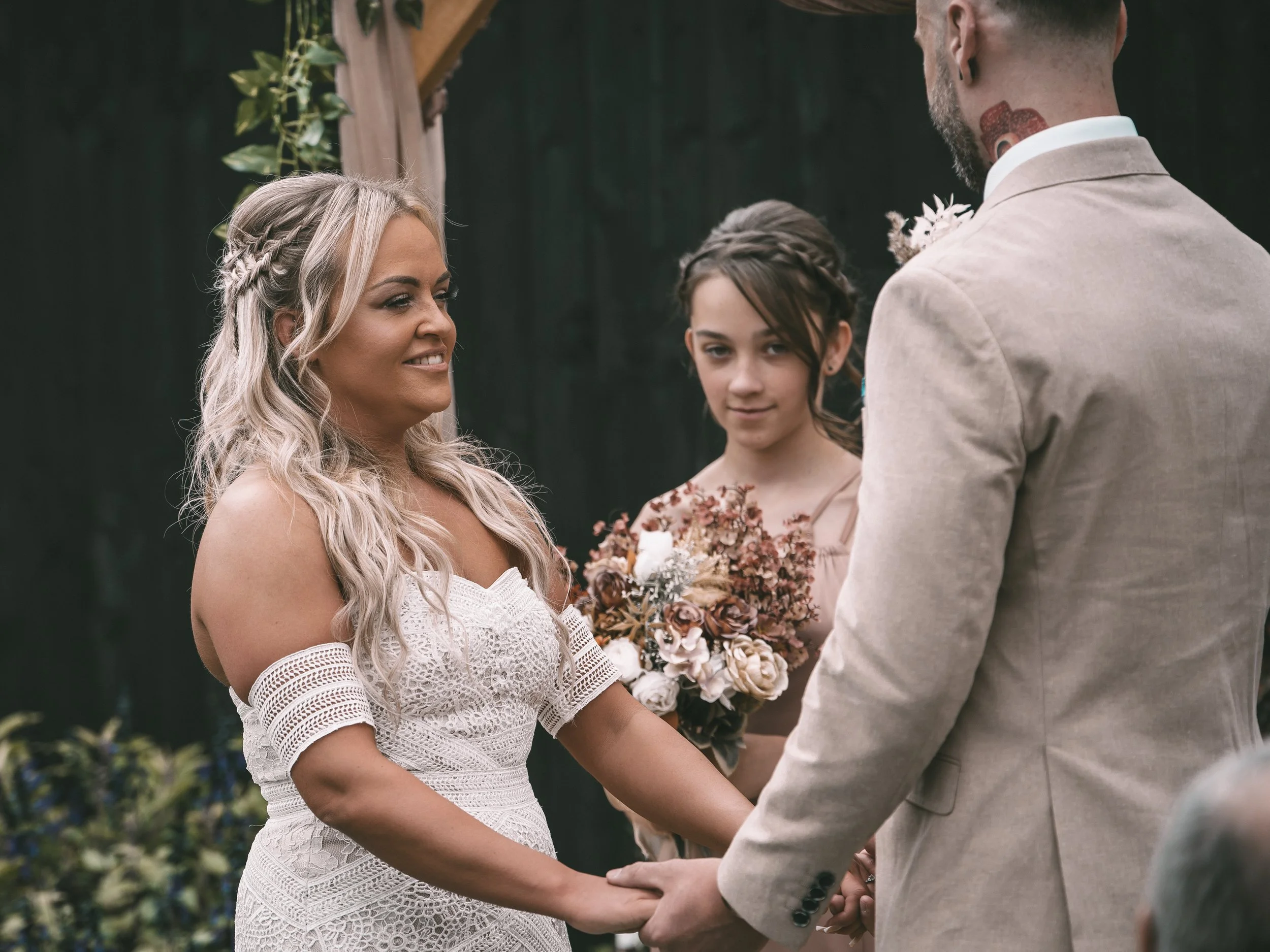 A bride and groom hold hands during their wedding ceremony, with a bridesmaid standing in the background holding a bouquet of flowers.
