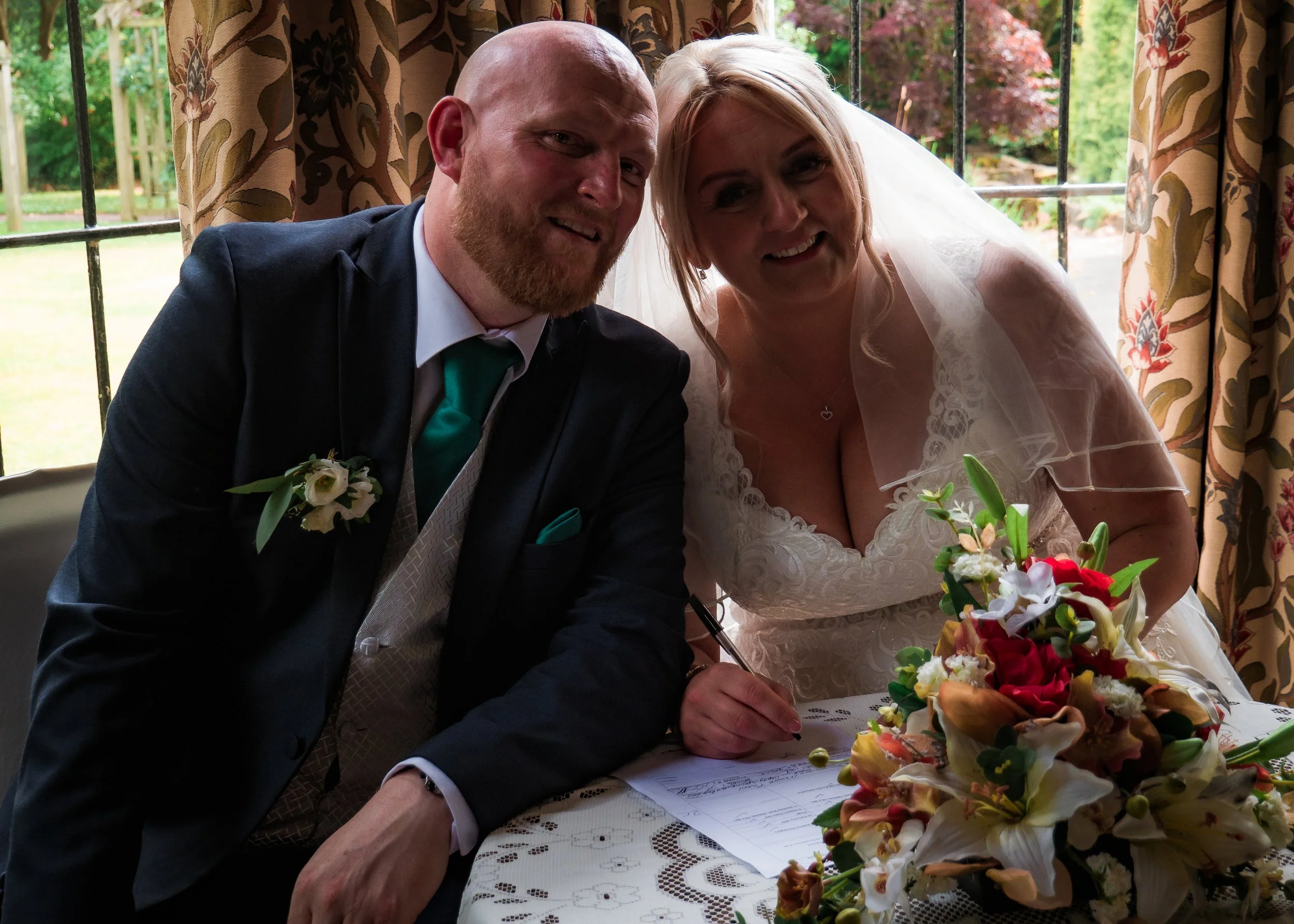 A newlywed couple is signing their marriage registry during a wedding ceremony. The groom is dressed in a dark suit with a teal tie and boutonniere, and the bride wears a white wedding dress with a veil. They are indoors, sitting at a table with a fl