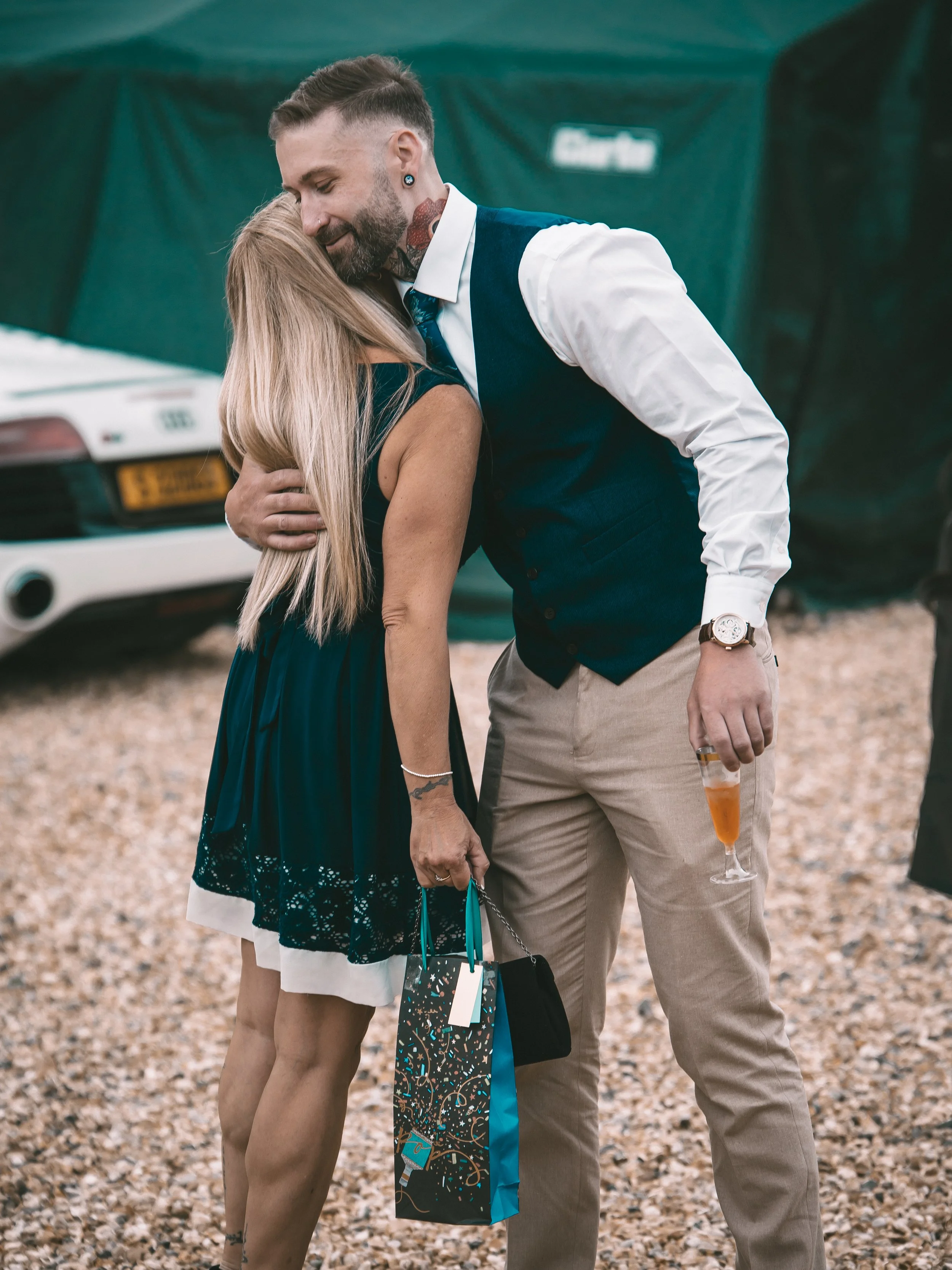 A man and woman embracing at a celebration, with the woman holding a gift bag and the man holding a cocktail glass.