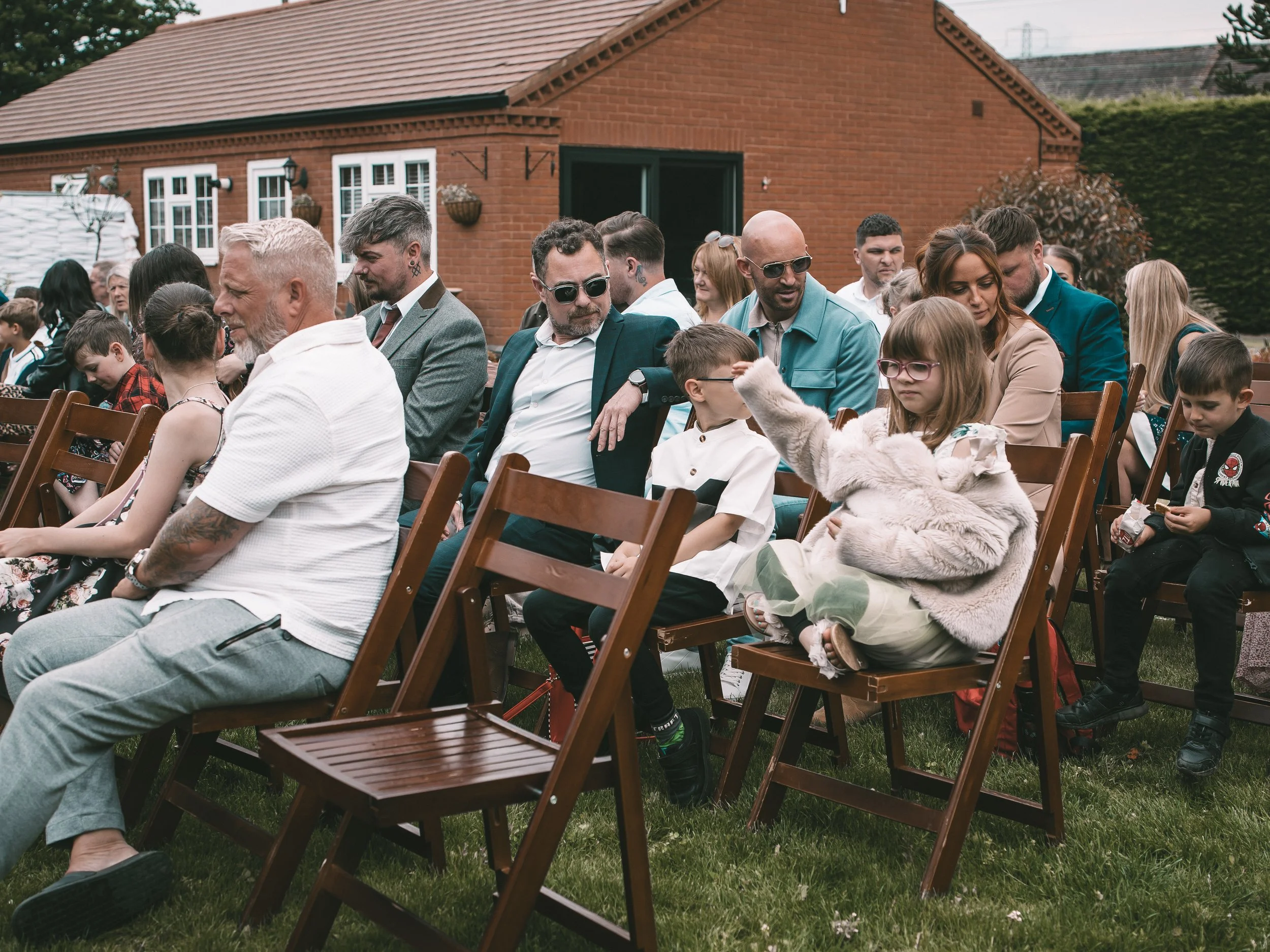 People sitting on wooden chairs outdoors during a gathering or event in front of a red brick house.