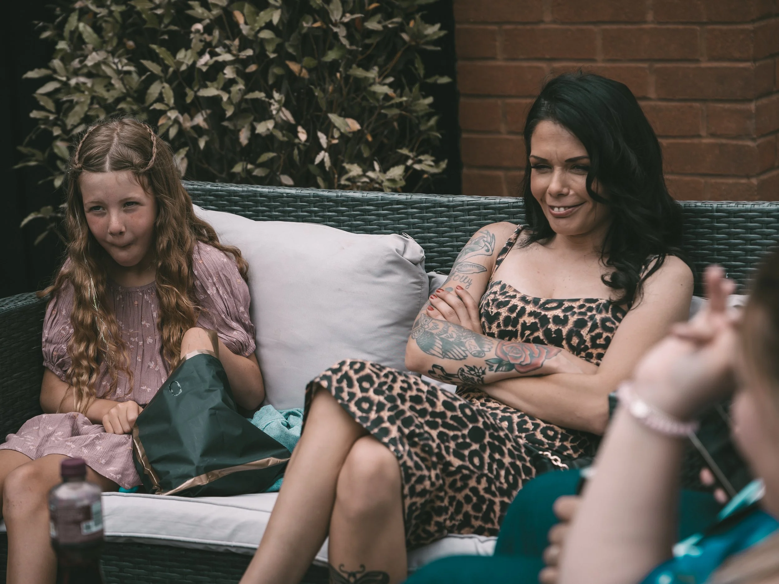 A young girl and a woman with tattoos sitting on a outdoor wicker couch, talking and smiling with a brick wall and foliage in the background.