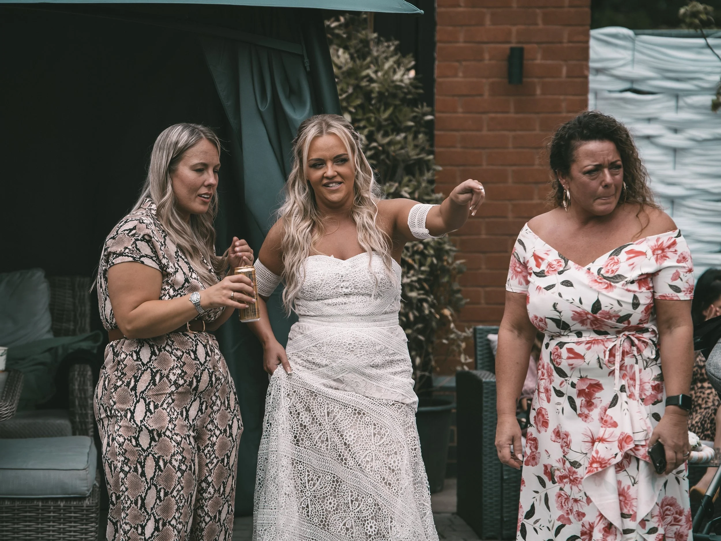 Three women standing outdoors, two women facing forward and one woman with blonde hair in a white crochet dress, with one arm extended, in front of a brick wall and outdoor furniture.